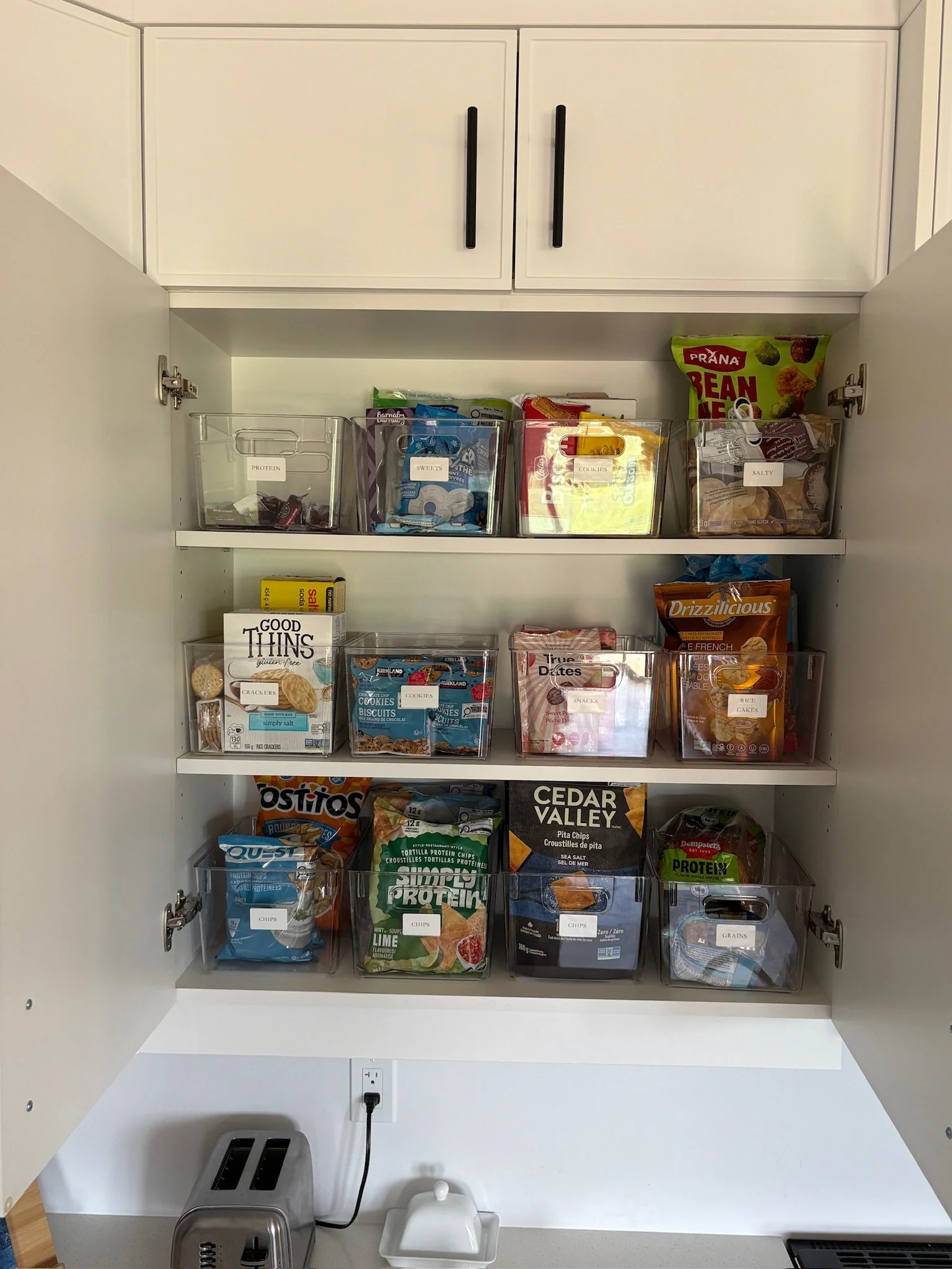 Open kitchen cabinet with clear storage bins labeled for protein, sweets, cookies, salty snacks, chips, rice cakes, and grains, containing various snack items, with a toaster and electric kettle on the white countertop below.