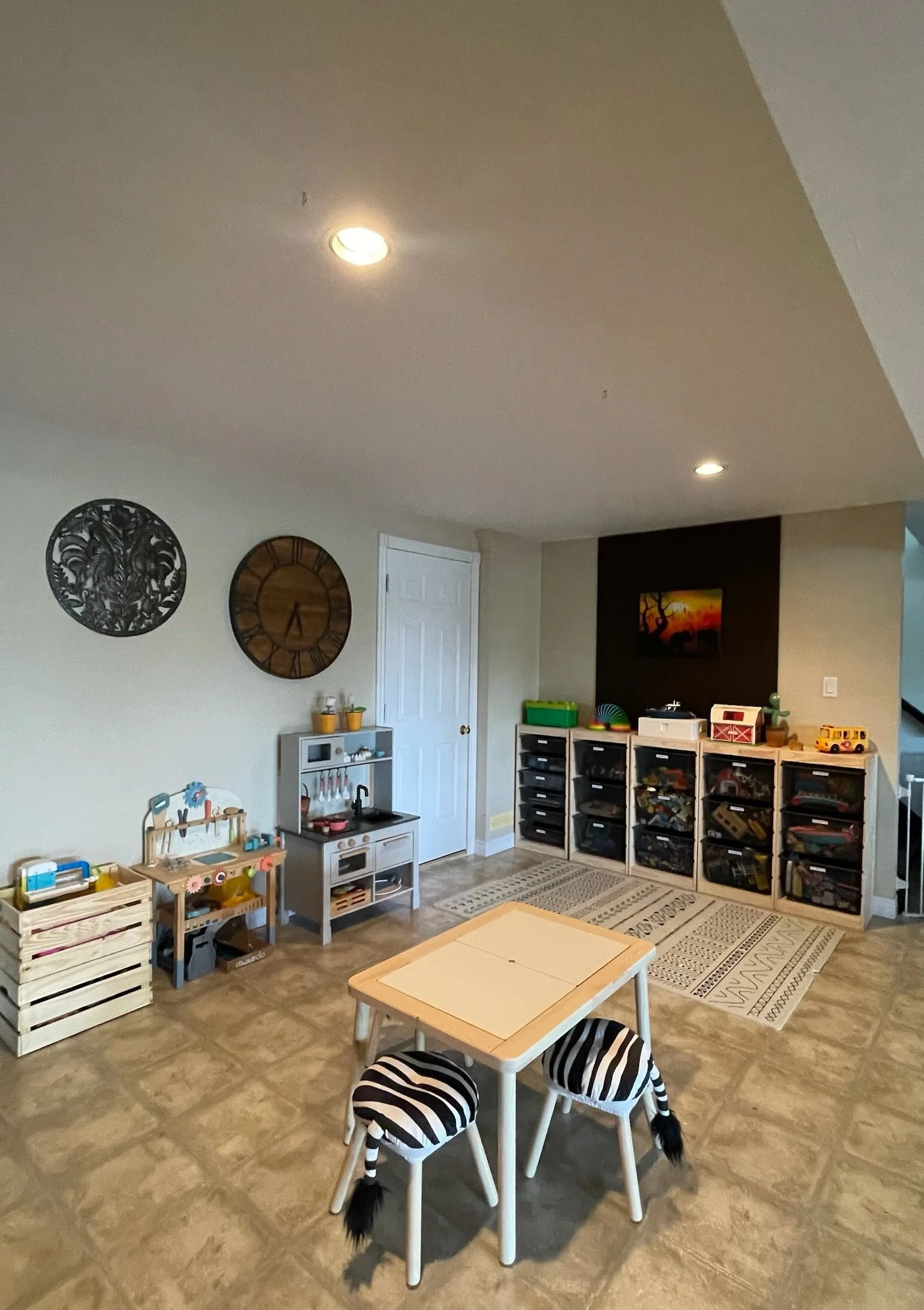 Kid's playroom with small table and two zebra-striped chairs, wall decor including a clock and decorative wall art, storage units with toys, and a rug on a tiled floor.