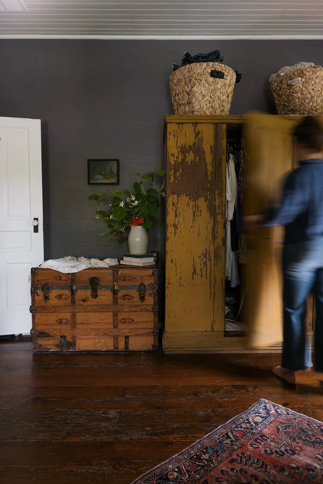 Photography of a moody blue interior design space with an old wood trunk, flowers in a grey vase, white door, yellow worn wordrobe with a person opening the door.