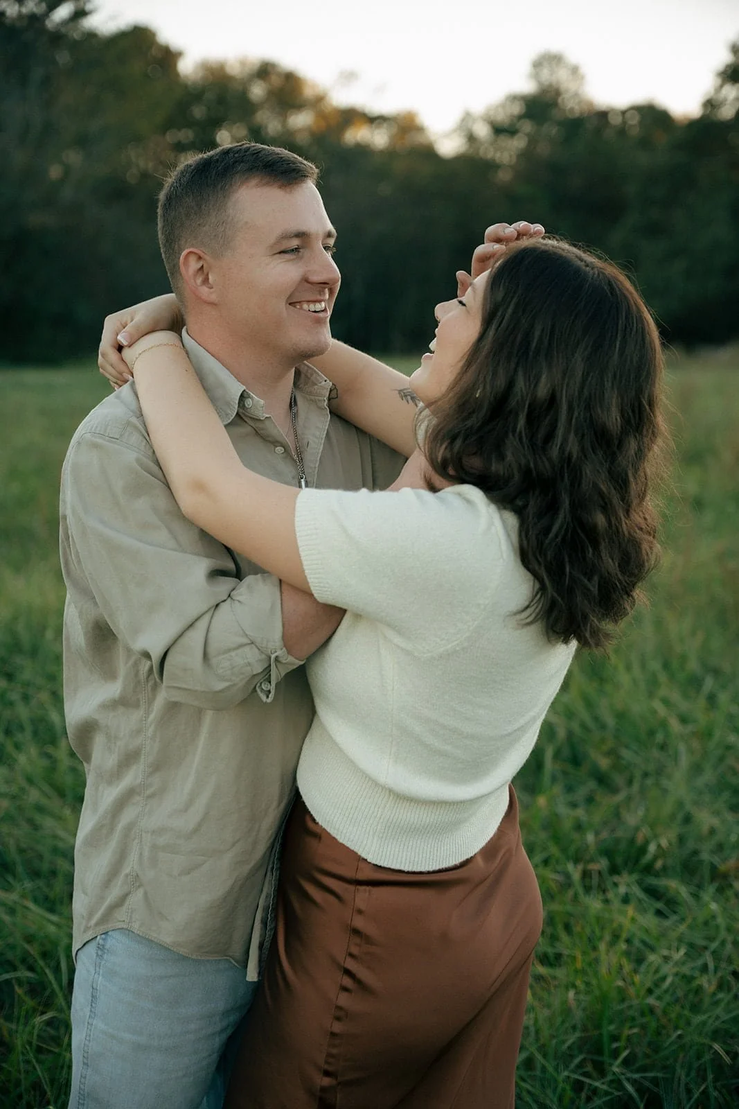 man and woman in field embracing. woman has arms around man's neck with eyes closed laughing. man brushing woman's hair out of eyes