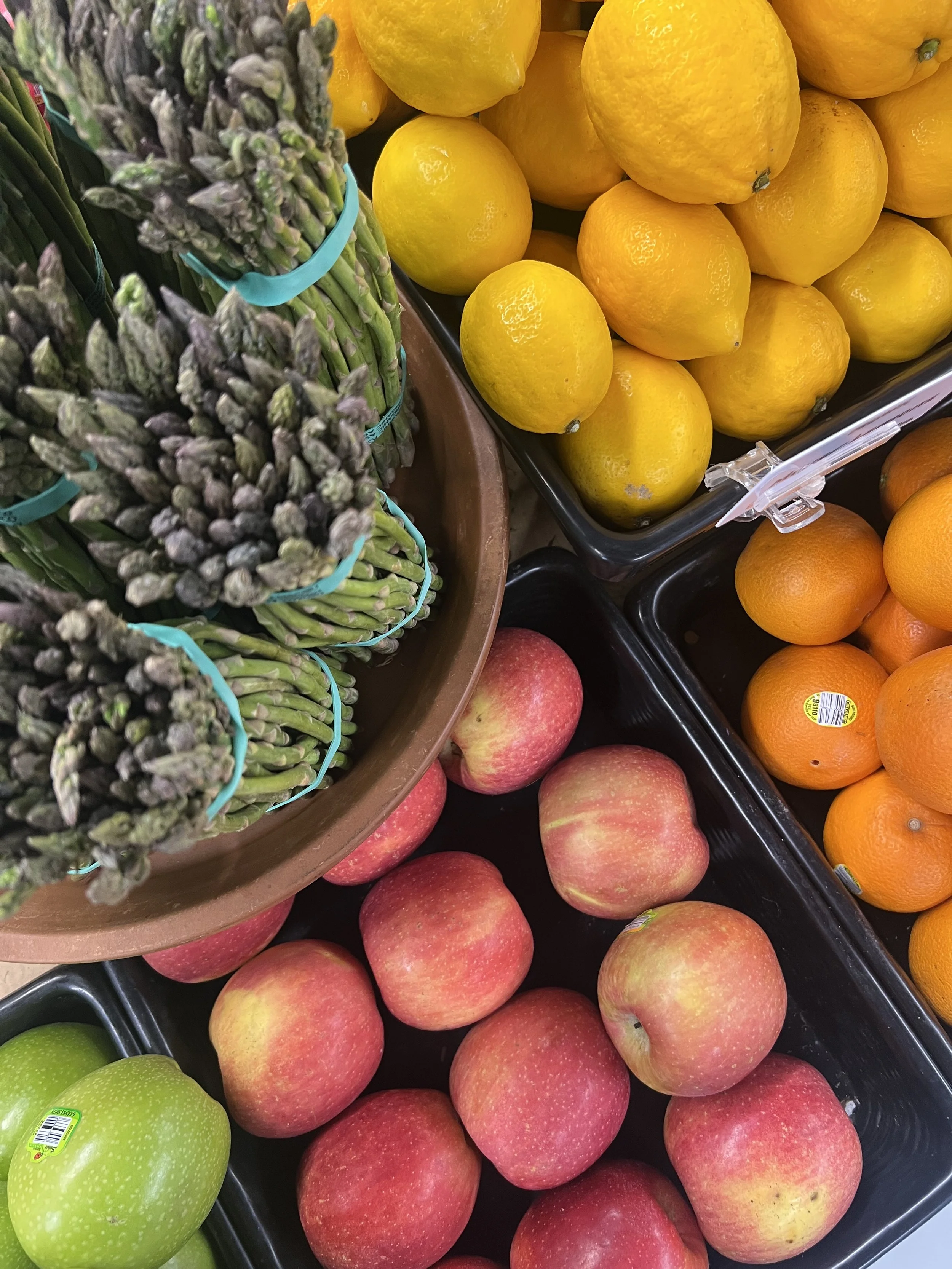 Bunches of fresh green asparagus in plastic bands, yellow lemons, orange oranges, and red apples displayed in a grocery store.