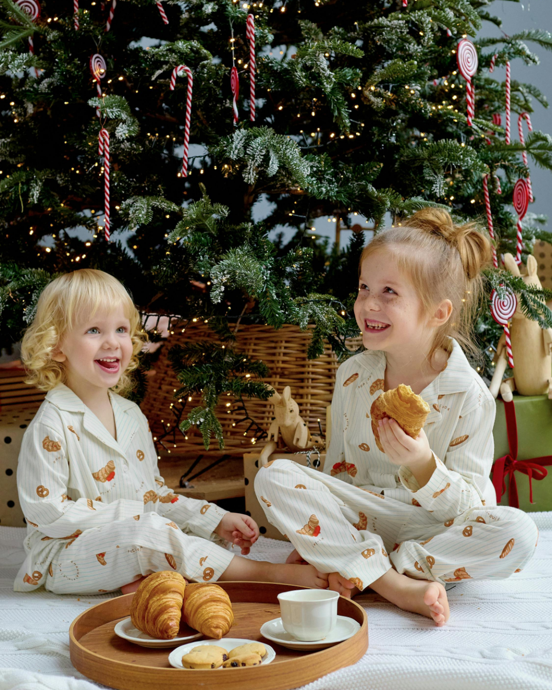 two sisters in matching Christmas pajamas eating snacks, croissants and cookies, under a Christmas tree. Tree has a rattan base cover.
