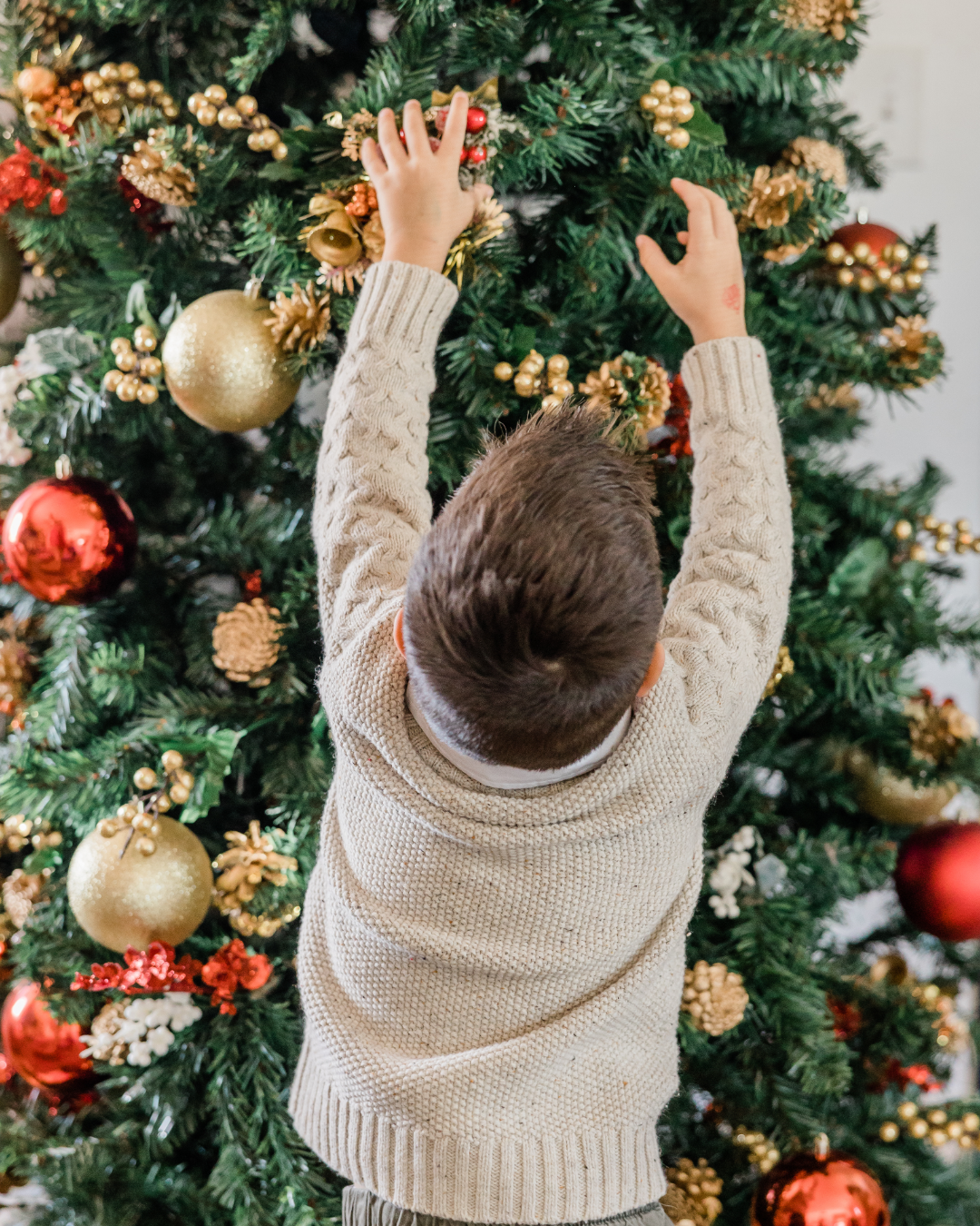 Little Boy decorating Christmas tree with pinecones and berries, bells and ornaments in gold and red