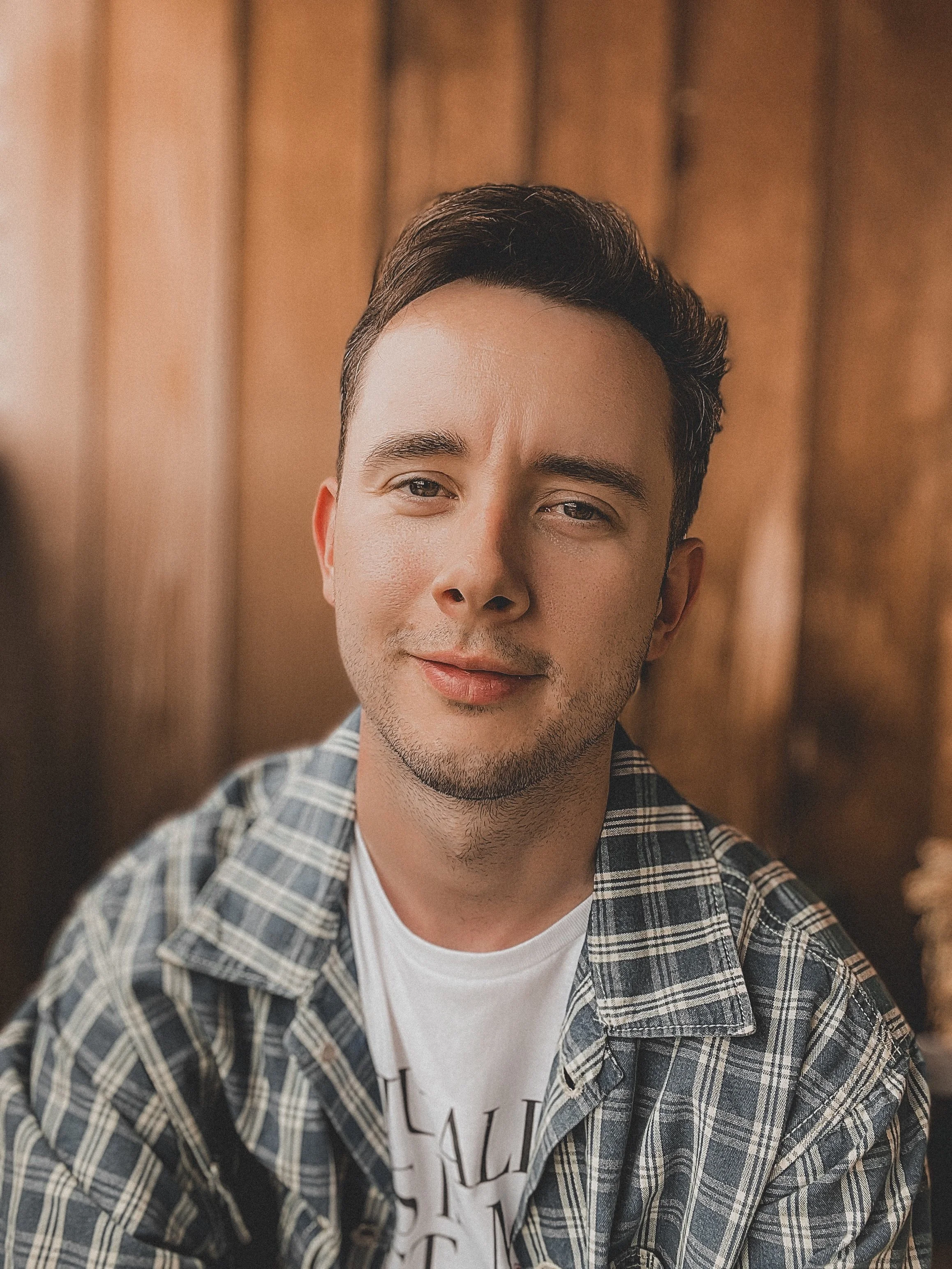 A man wears a denim jacket and a white t-shirt. He looks at the camera and smiles. He is on a background of wood panelling.