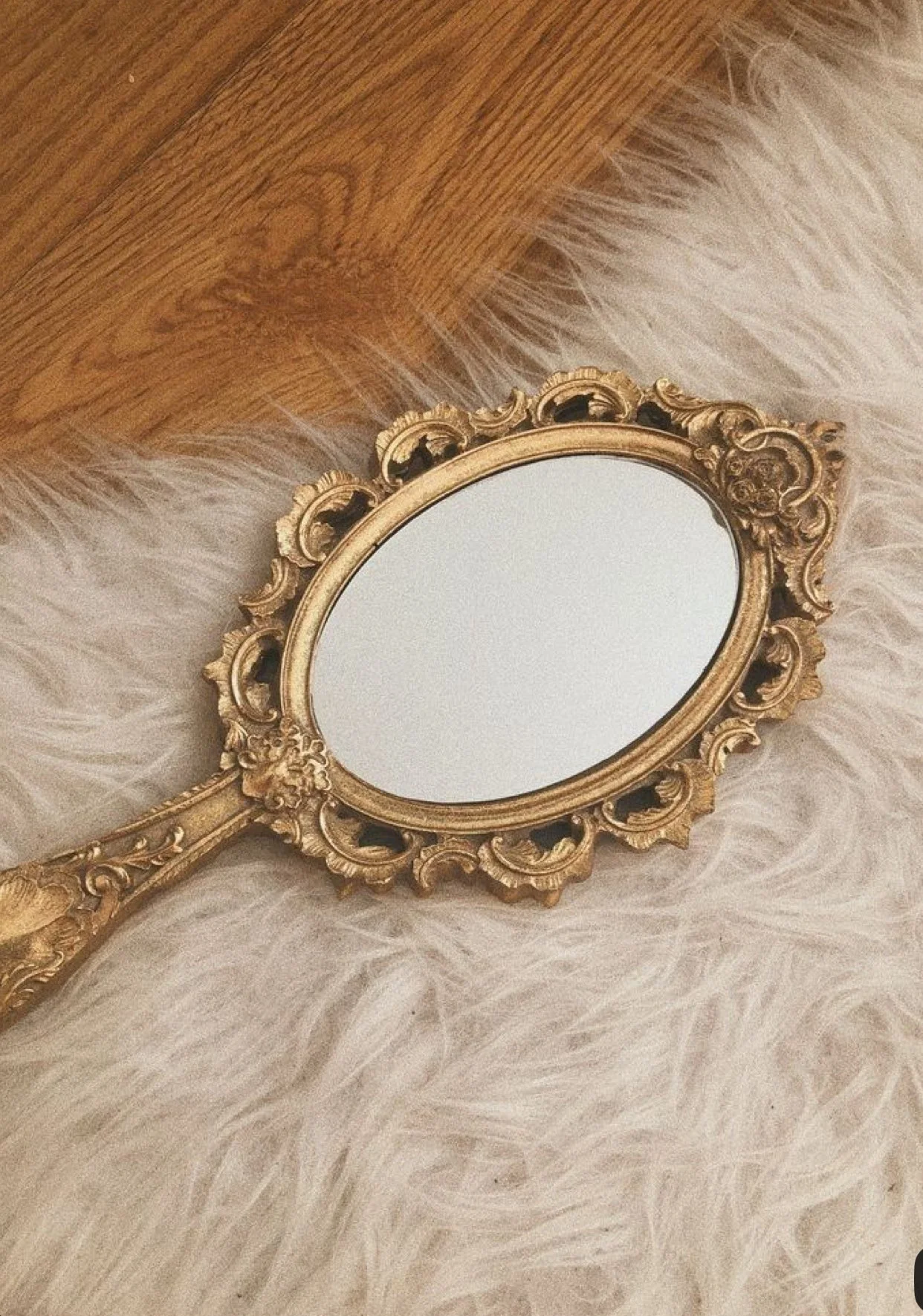 Un miroir ancien avec un cadre doré et orné, posé sur un tapis blanc et une surface en bois.