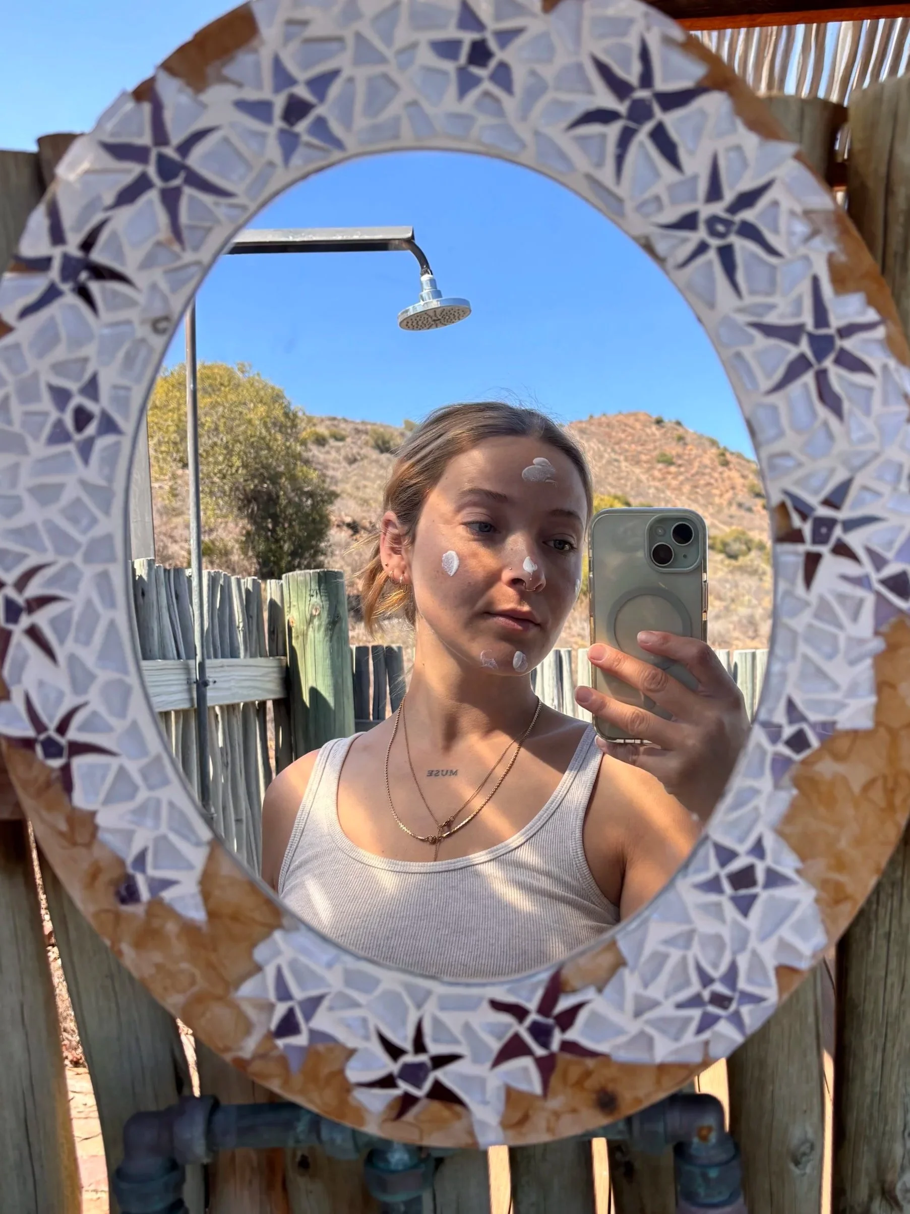 Jeune femme se prenant en selfie dans un miroir, en extérieur, avec une montagne en fond. Elle porte un débardeur blanc, deux colliers, et a de la crème ou du maquillage blanc sur le visage.