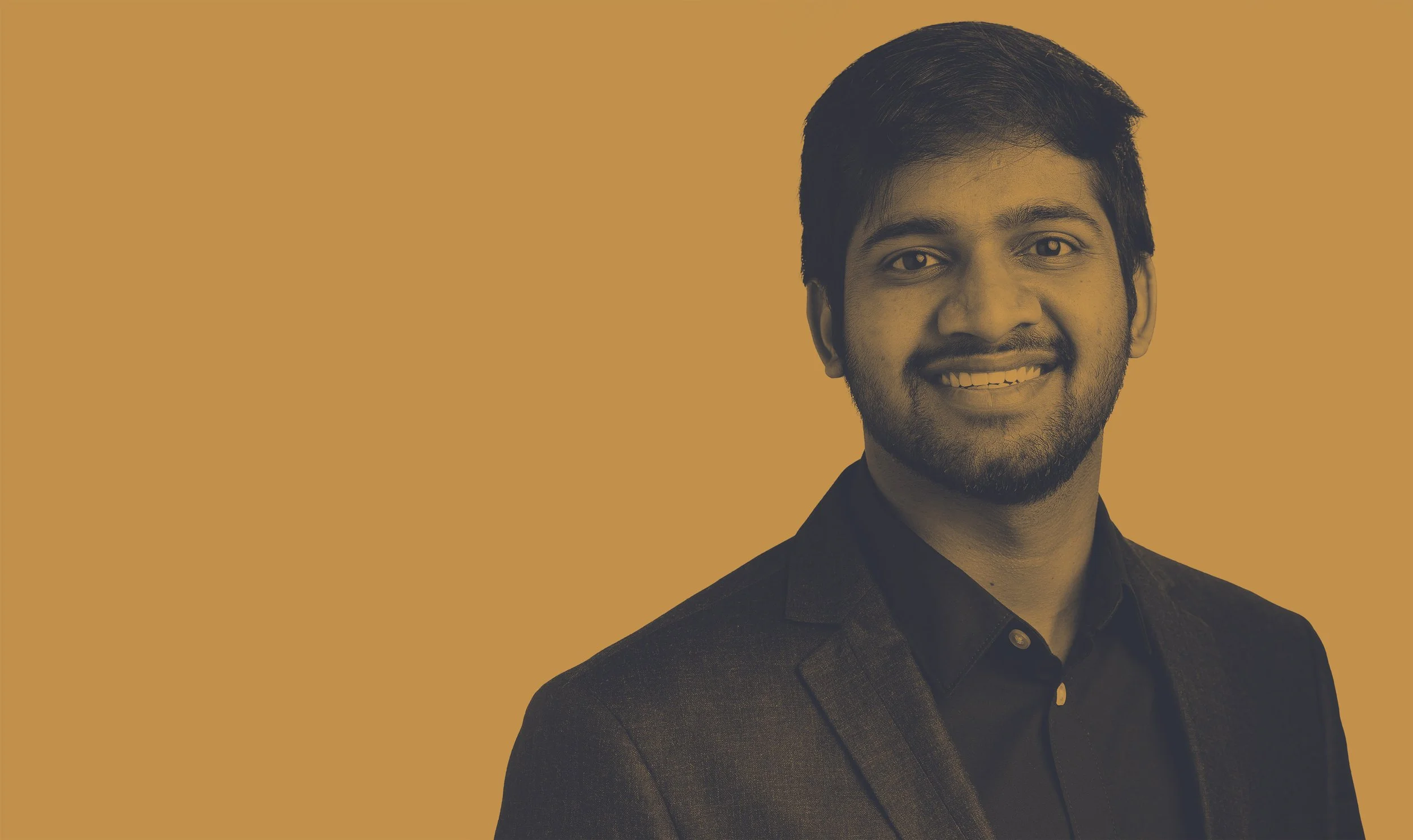 Portrait of a young man with a beard and short dark hair, smiling, in a dark blazer and shirt, against a solid mustard-colored background.