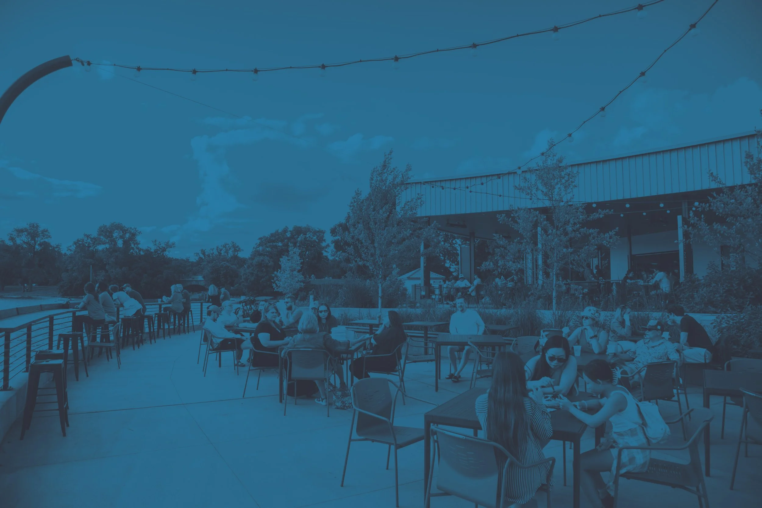 Outdoor patio with people sitting at tables and a bar, some people are reading, talking, and using their phones under string lights on a sunny day with trees and a building background.