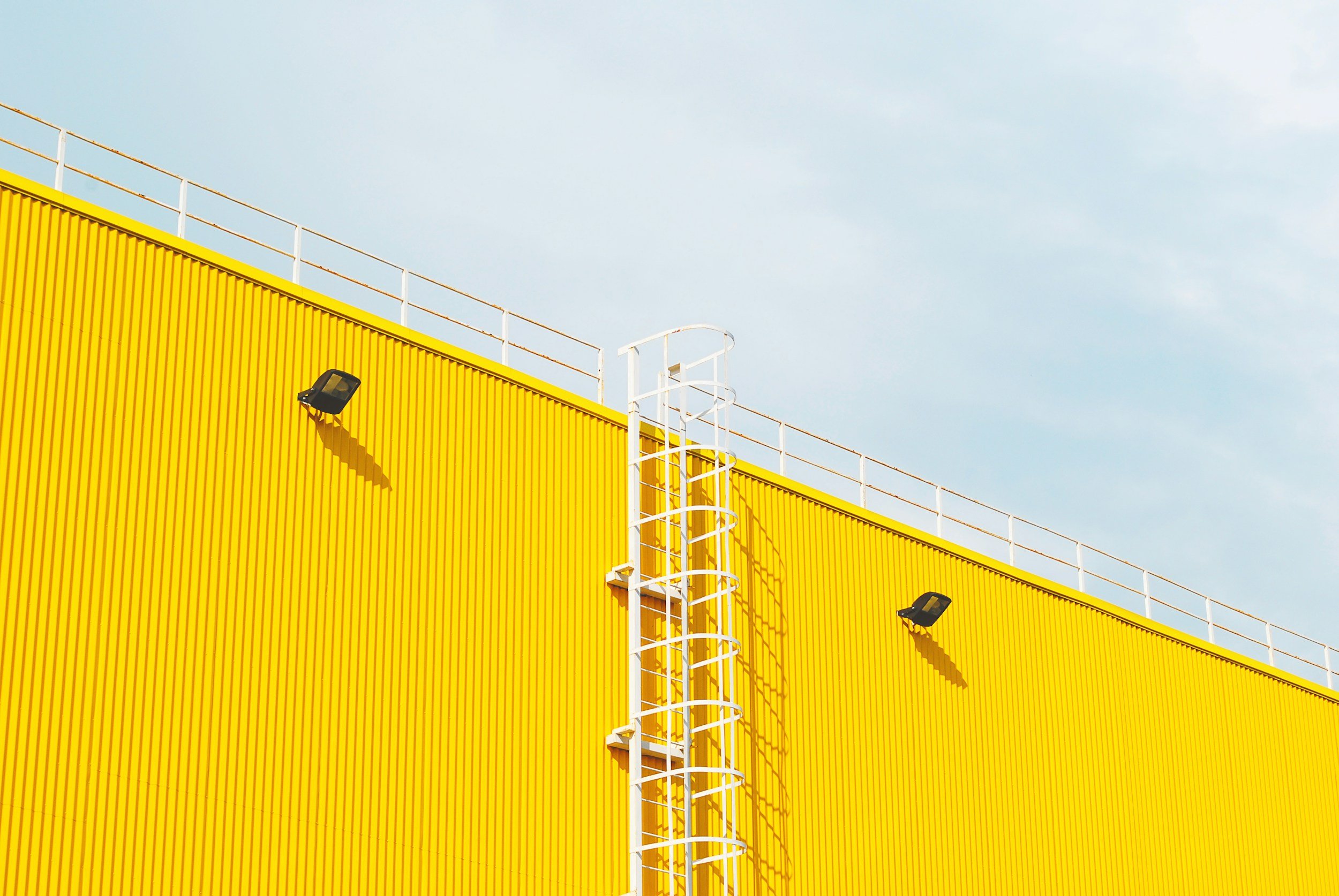 A yellow building with vertical ridges, white railing on top, a white fire escape ladder, and two black outdoor lights against a cloudy sky.