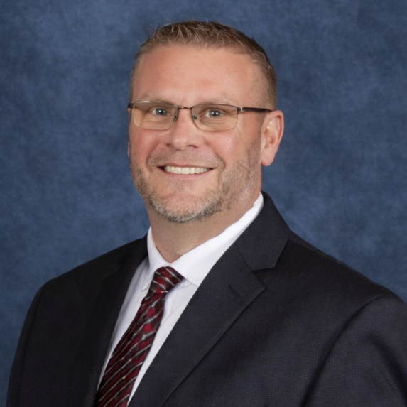 Professional man wearing glasses, a black suit, white shirt, and patterned tie, smiling against a blue textured background.