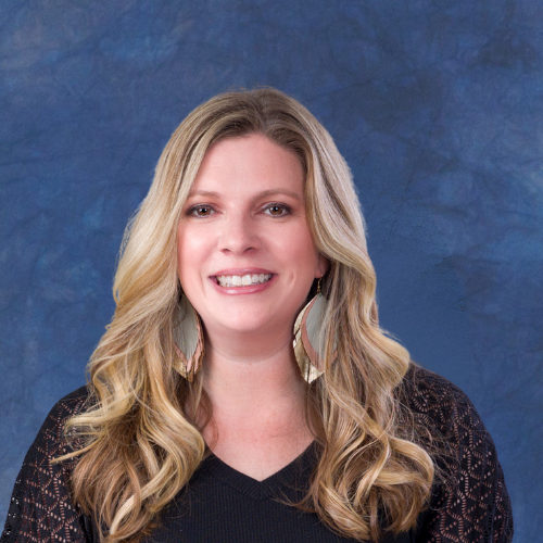 Portrait of a woman with blonde wavy hair and light makeup, wearing a patterned top with yellow, black, and white colors, and pearl earrings, against a blue textured background.