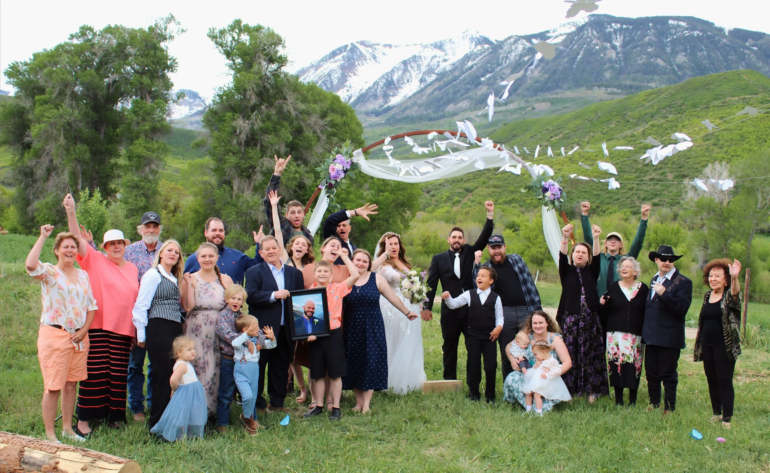 A wedding celebration outdoors with a large group of people standing under a decorated arch, surrounded by green hills and mountains in the background. The group is smiling and raising their hands in celebration.