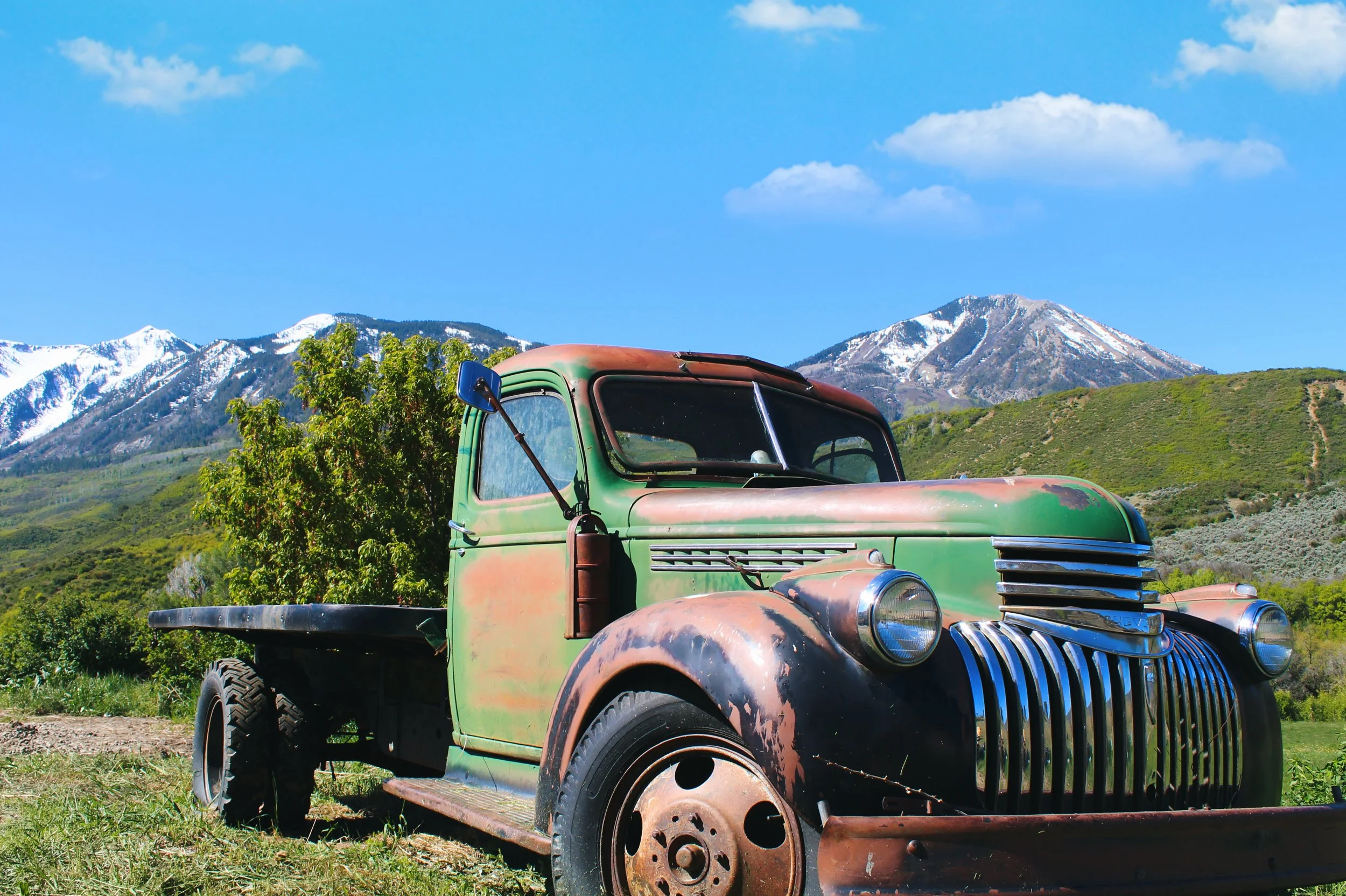 An old, rusty green truck with faded paint, parked on grass with mountains and a clear blue sky with clouds in the background.