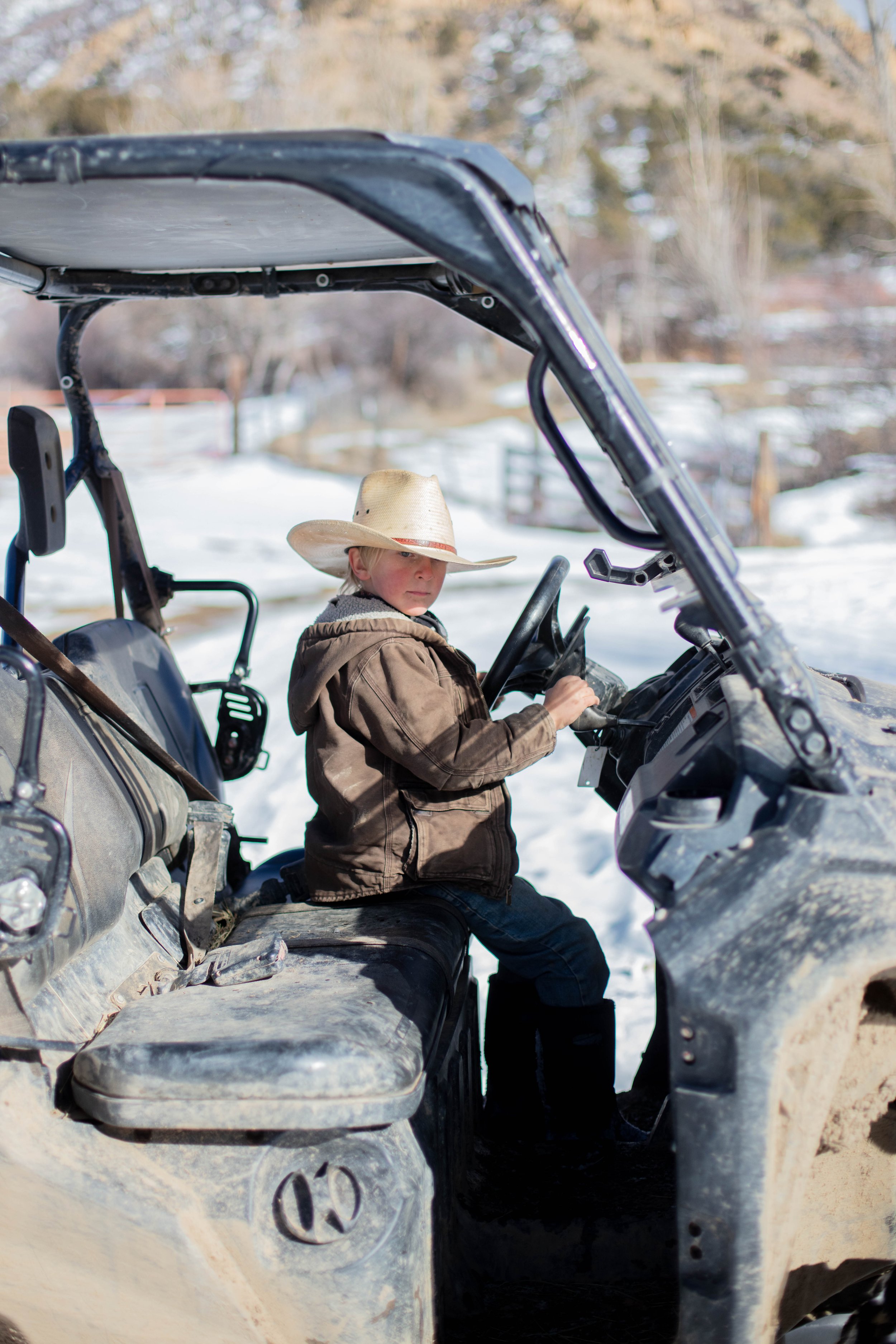 A young boy wearing a cowboy hat and brown jacket sitting on a utility ATV in a snowy outdoor area with trees in the background.