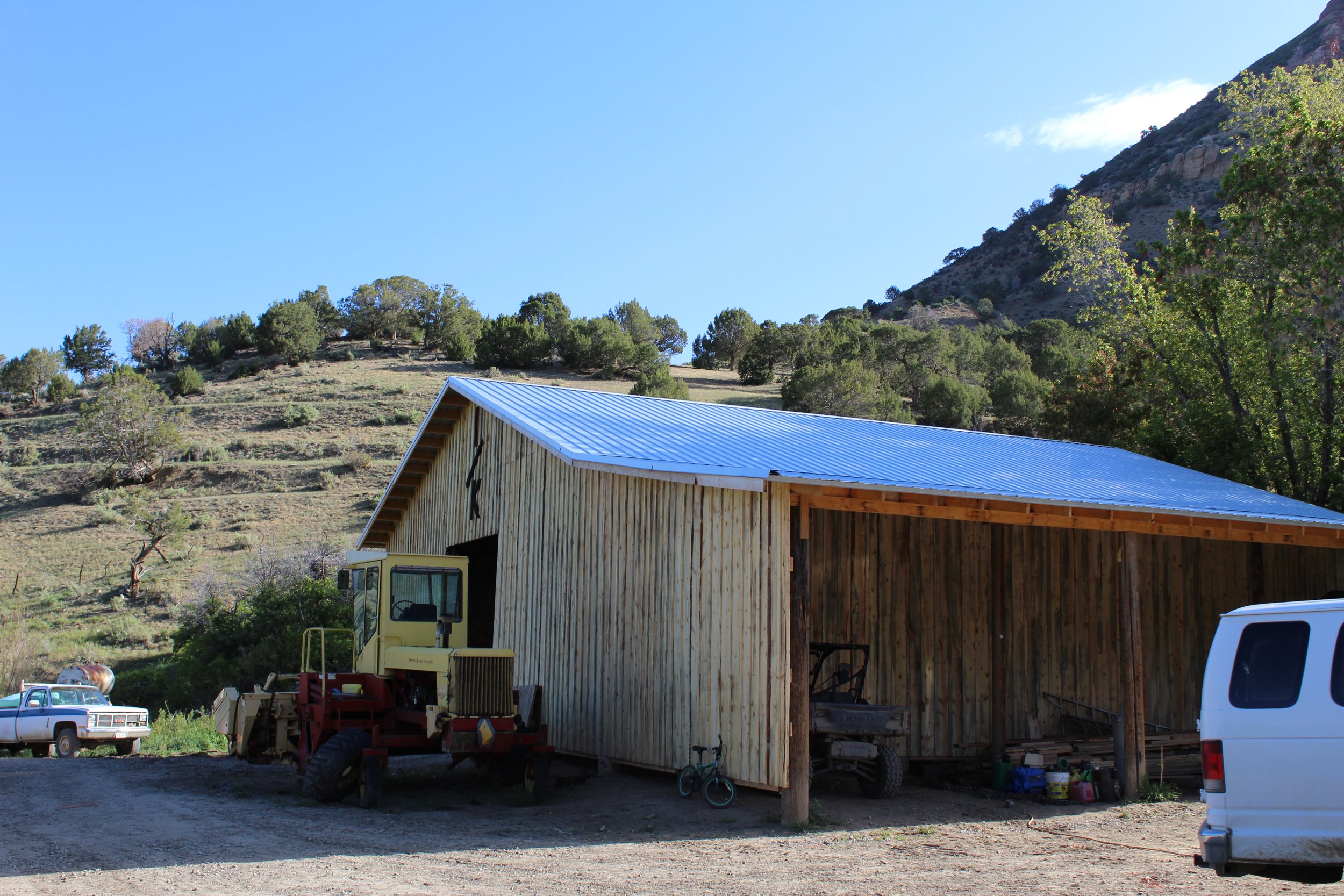 A rural scene with a wooden barn with a metal roof, farm equipment, and vehicles parked nearby, set against a backdrop of a hillside covered with trees and shrubs under a clear blue sky.