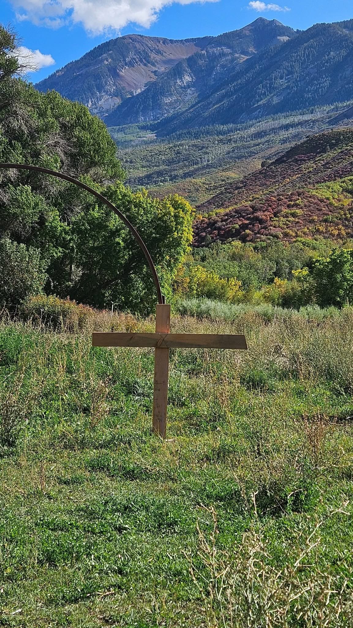 A wooden cross in a grassy field with colorful trees, mountains, and a blue sky in the background.