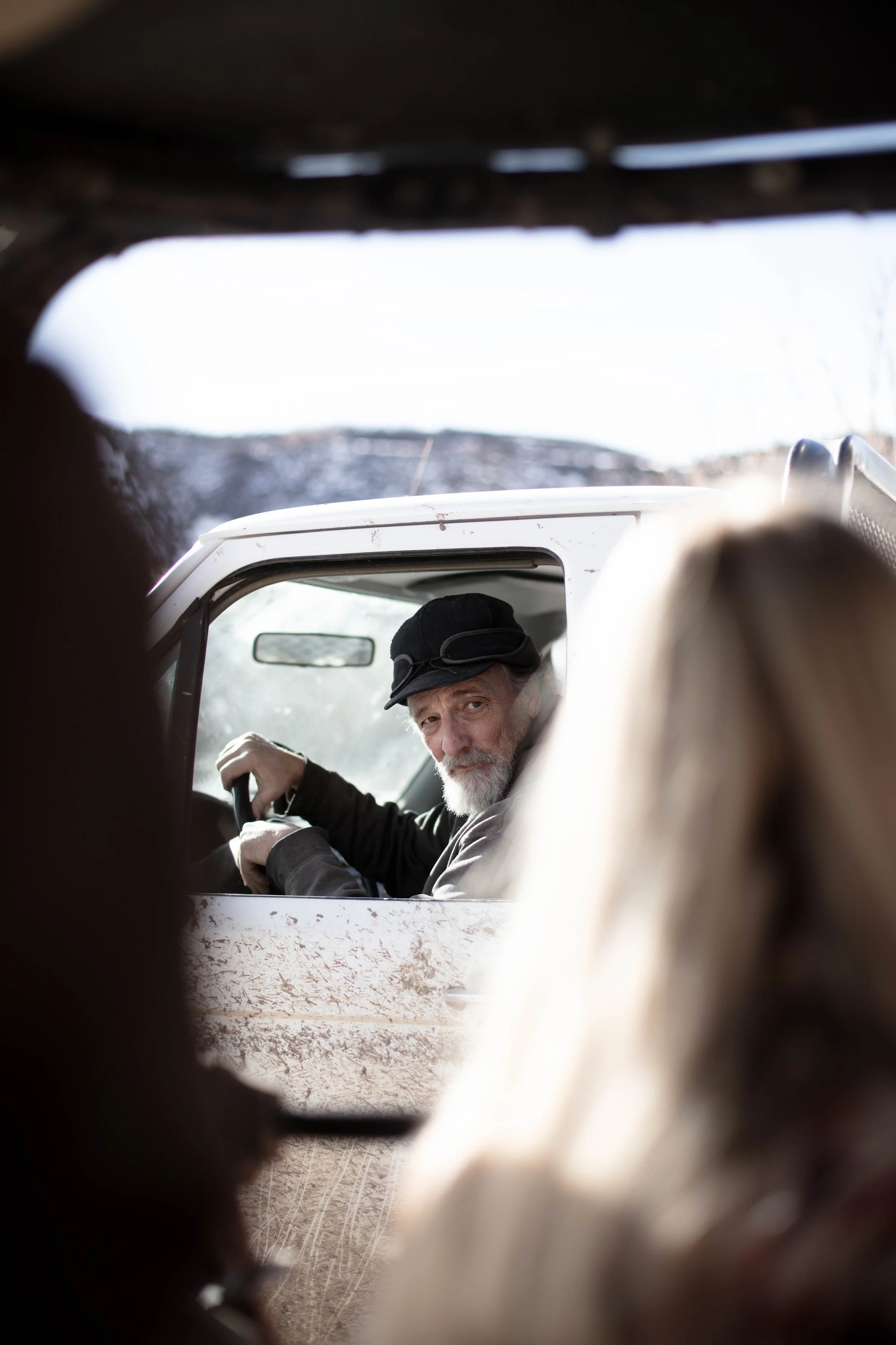 A man with a gray beard and black hat sitting in a white vehicle, looking through an opening in a dirt-covered structure, with blurred figures in the foreground.