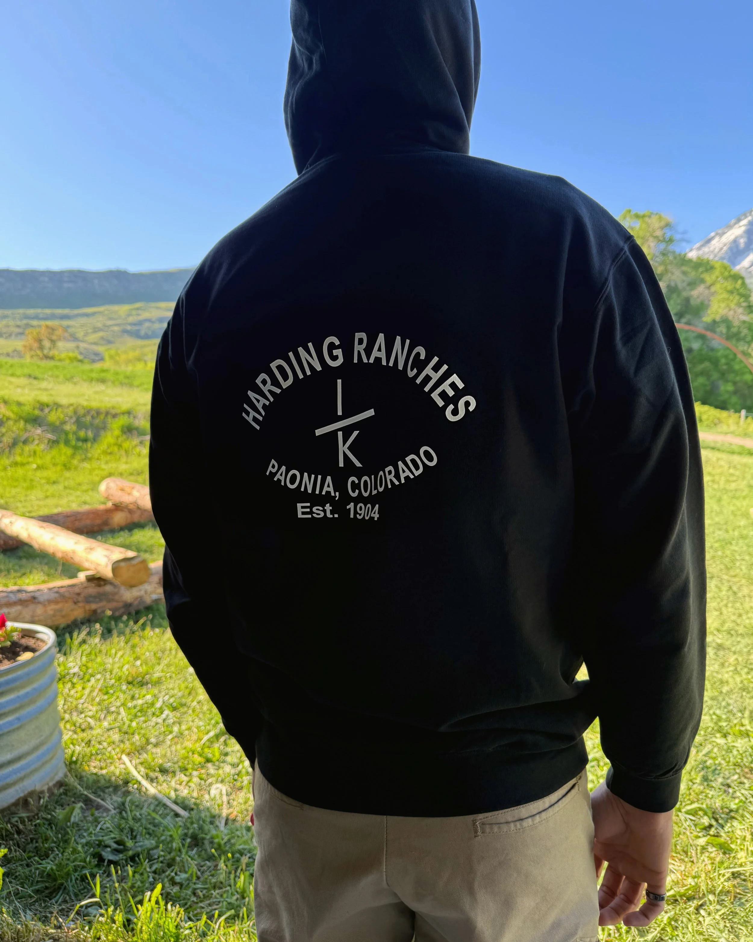 Person wearing a black hoodie with the words 'HARDING RANCHES, PAONIA, COLORADO, Est. 1904' on the back, standing outdoors on grass with mountains and trees in the background, under a clear blue sky.
