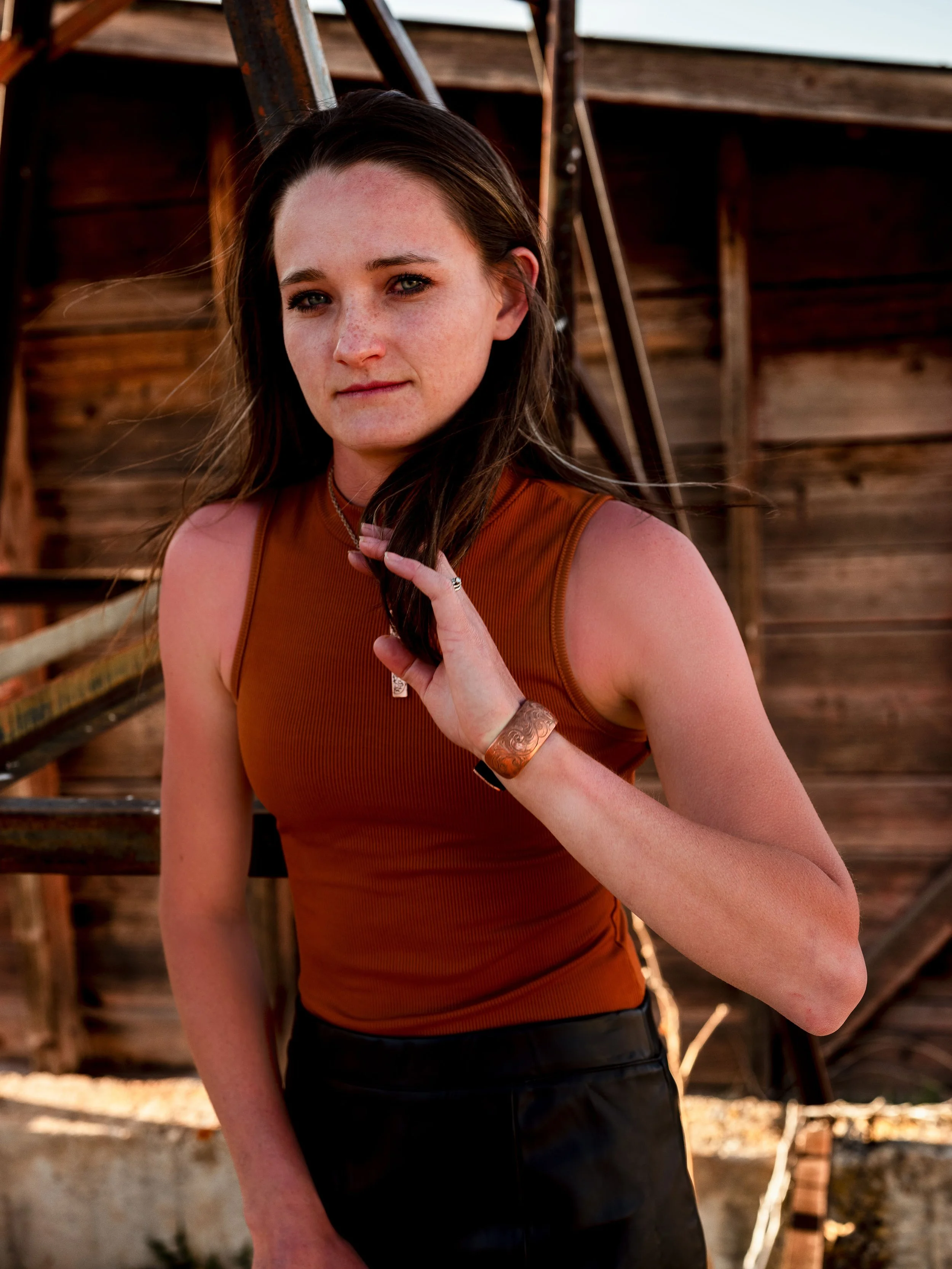A young woman with long dark hair and light skin, wearing a sleeveless rust-colored top and jewelry, outdoors near wooden stairs and structure, looking at the camera.