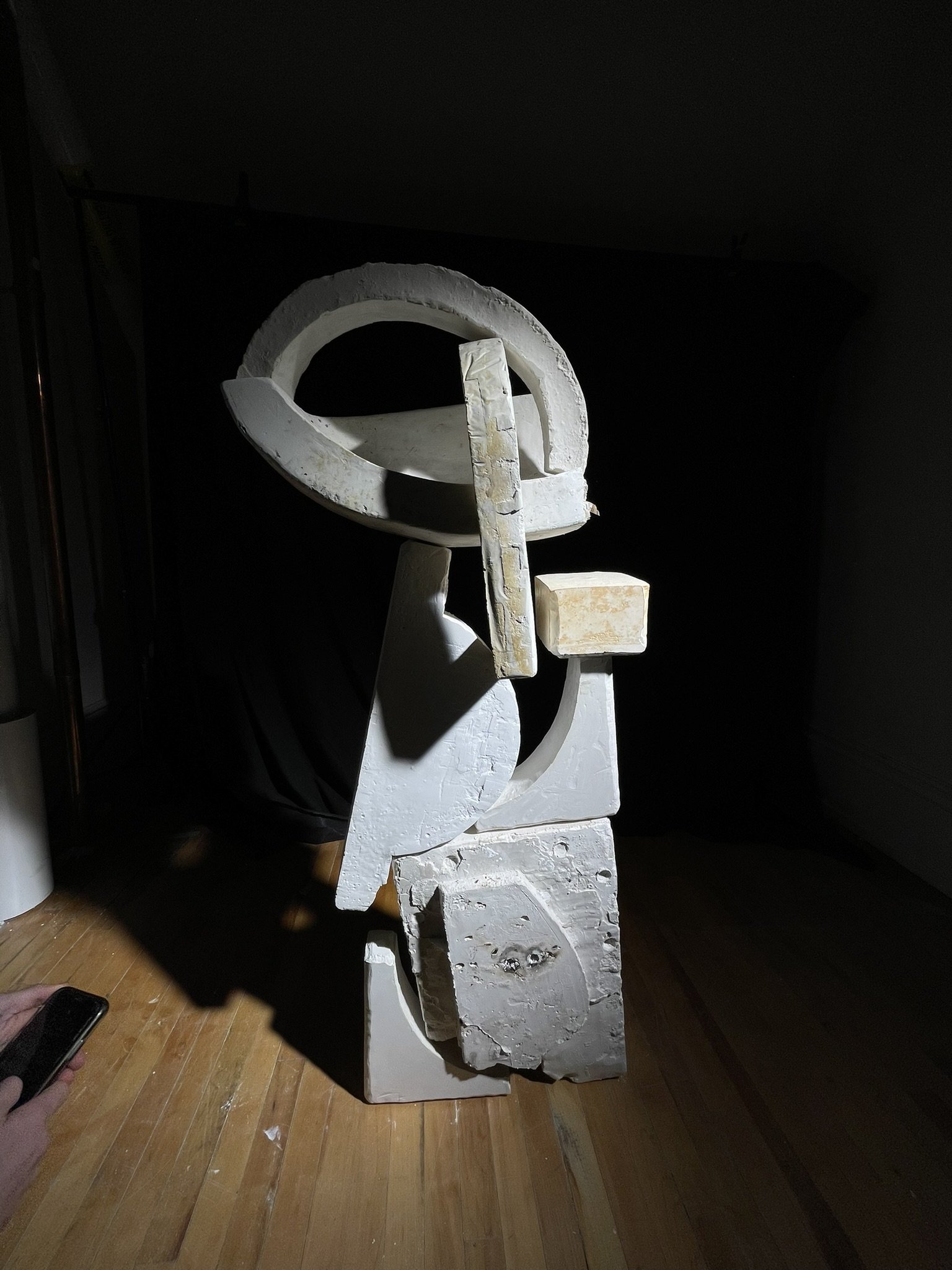 A white sculpture or art piece with abstract shapes and curves, standing on a wooden floor against a dark background.