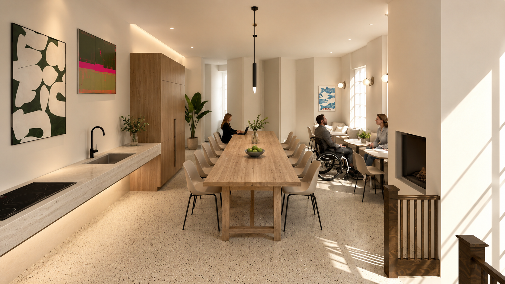 A spacious, modern communal dining and lounge area with a long wooden table, beige chairs, and artwork on the walls. There is a kitchen counter with a sink and stovetop on the left, and two people sitting at a small table on the right, one in a wheel