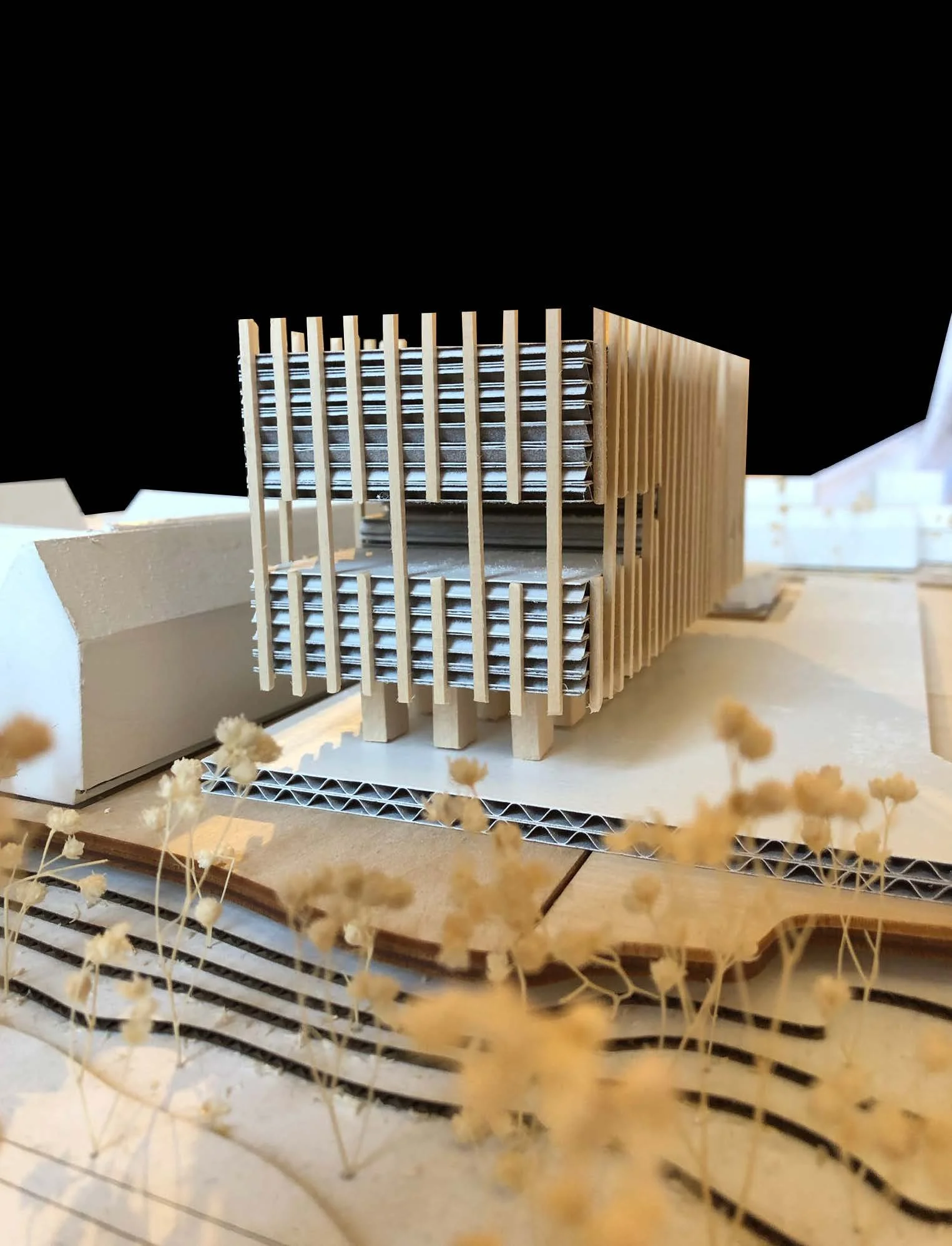 A detailed architectural model of a modern building made of wood and cardboard, with vertical wooden slats and layered structural elements, set on a base with small decorative dried flowers in the foreground.