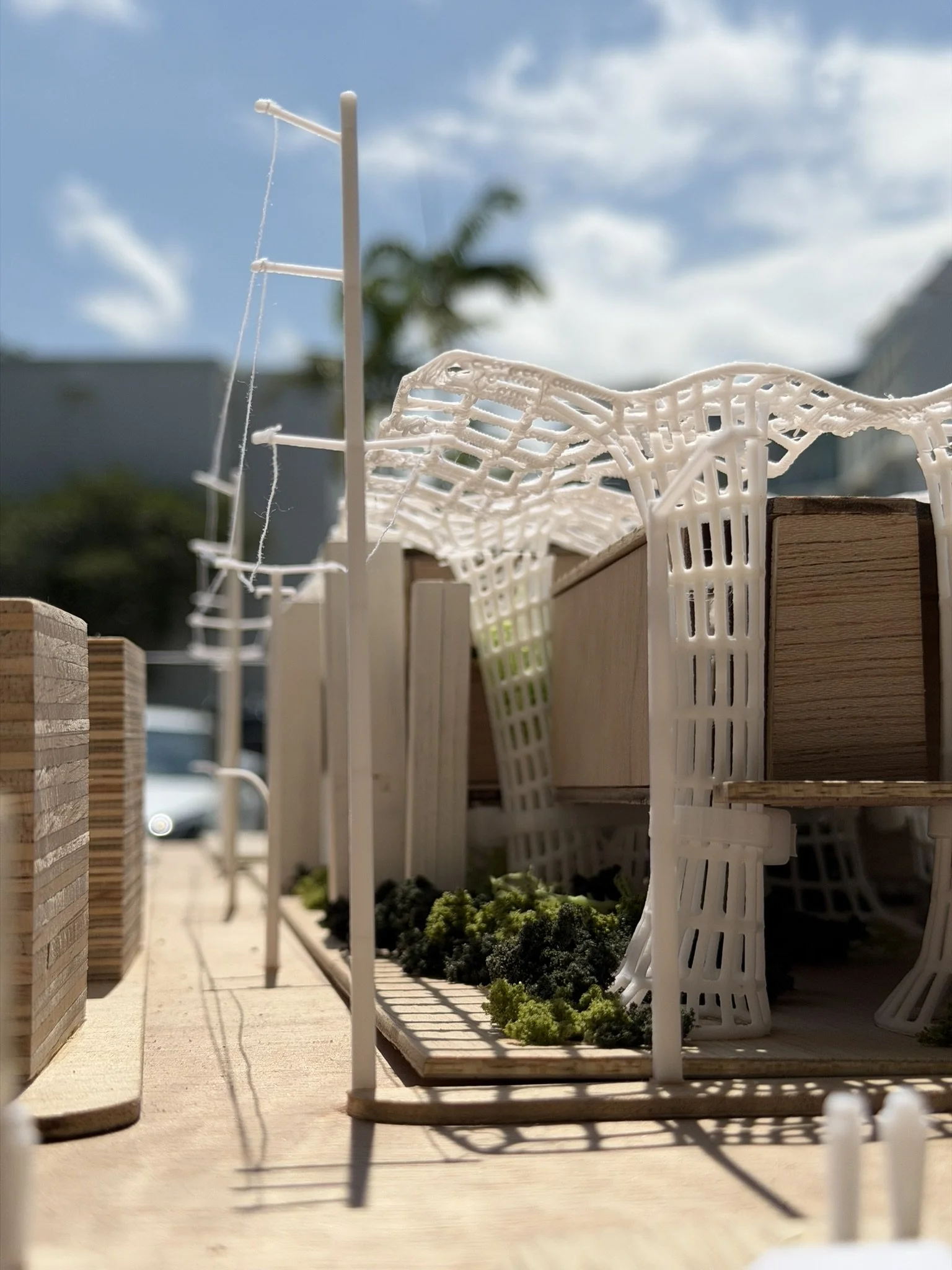 A detailed miniature architectural model of a modern building with a unique roof design, white support structures, small greenery, and a wooden base, set against a blue sky with clouds.