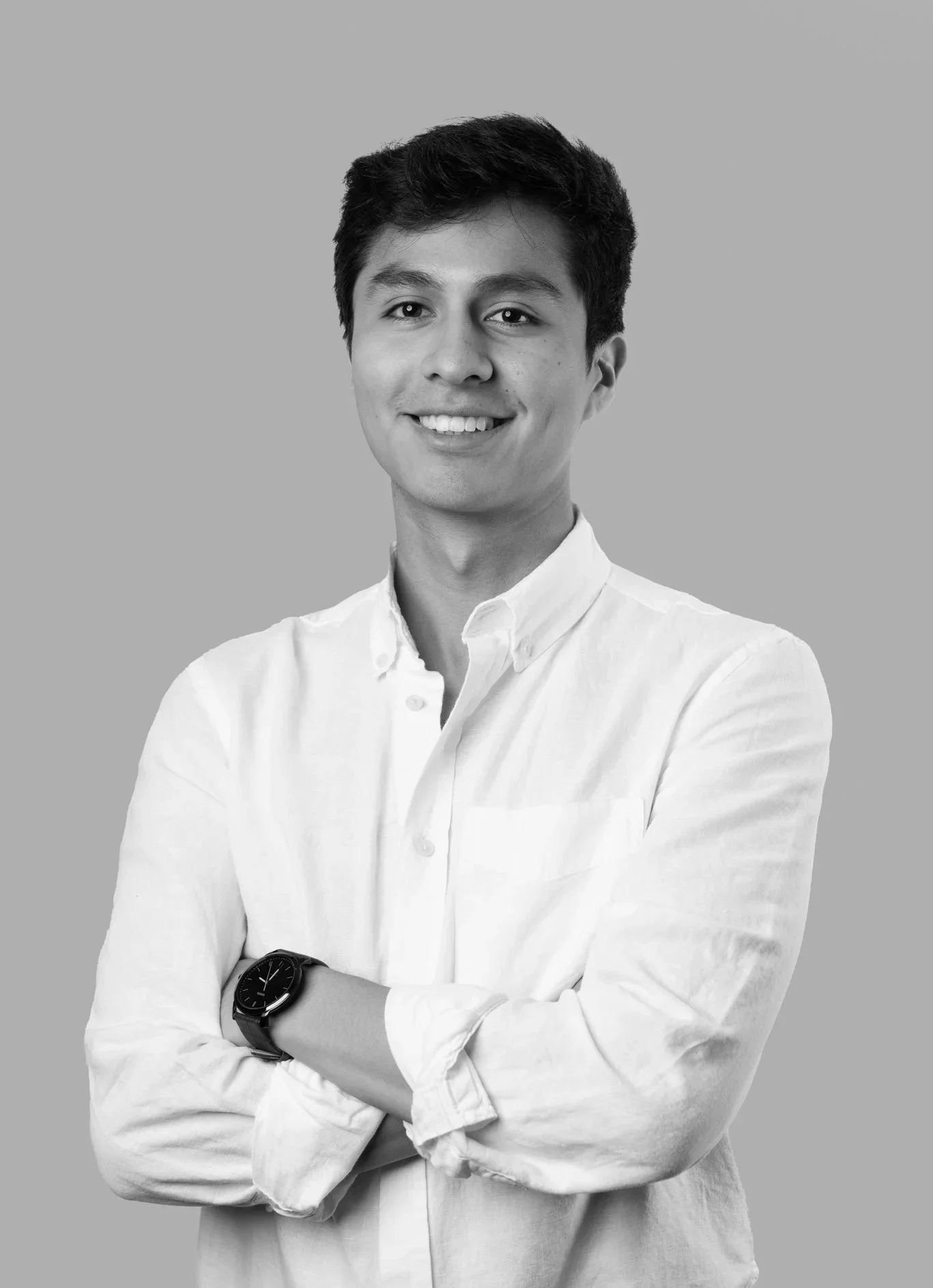 A young man with dark hair, smiling, wearing a white button-up shirt with arms crossed; wearing a black watch on his left wrist; on a plain light gray background.