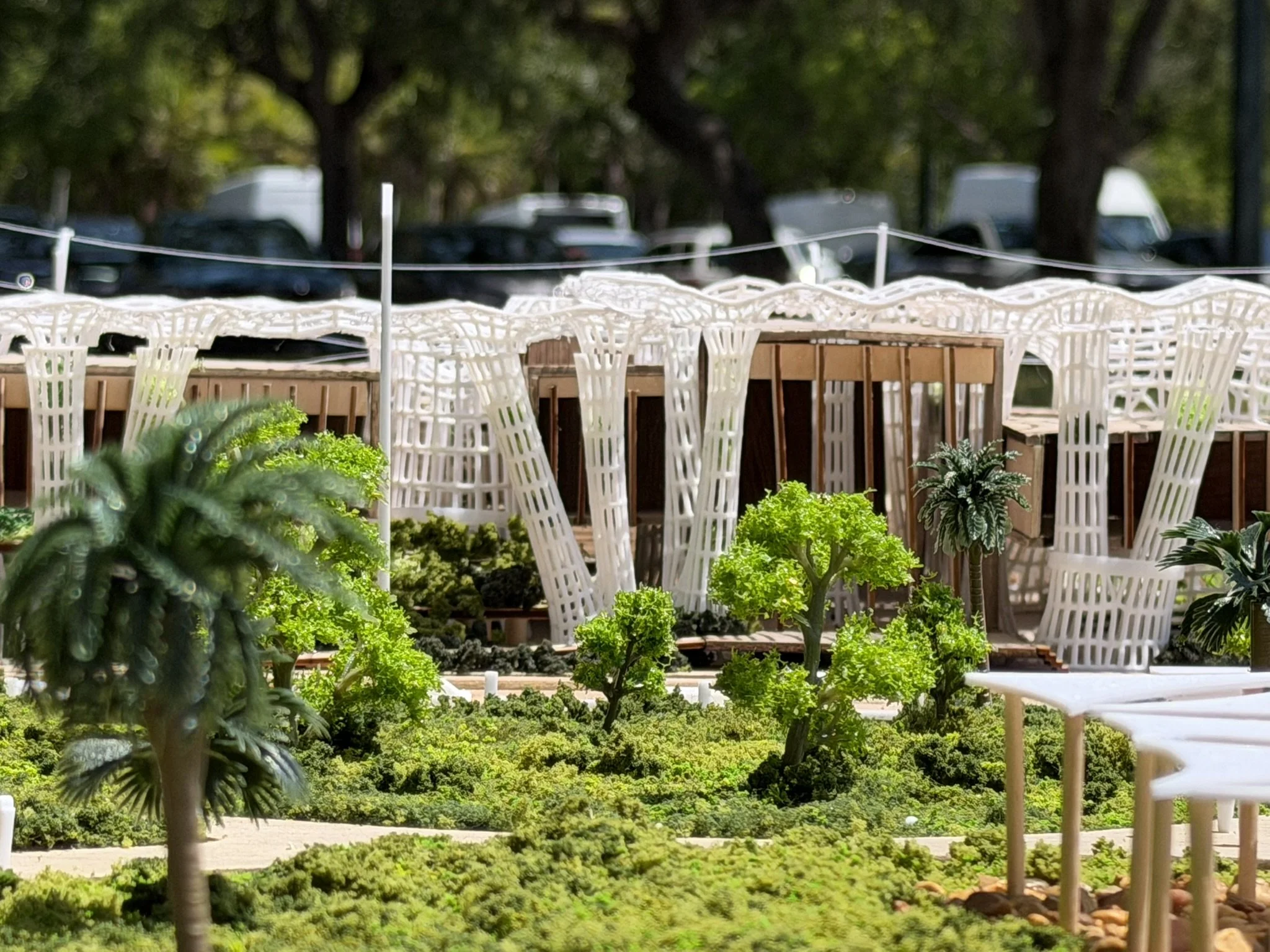 Outdoor scene with white chairs around a table, surrounded by green plants, trees, and parked cars in the background.