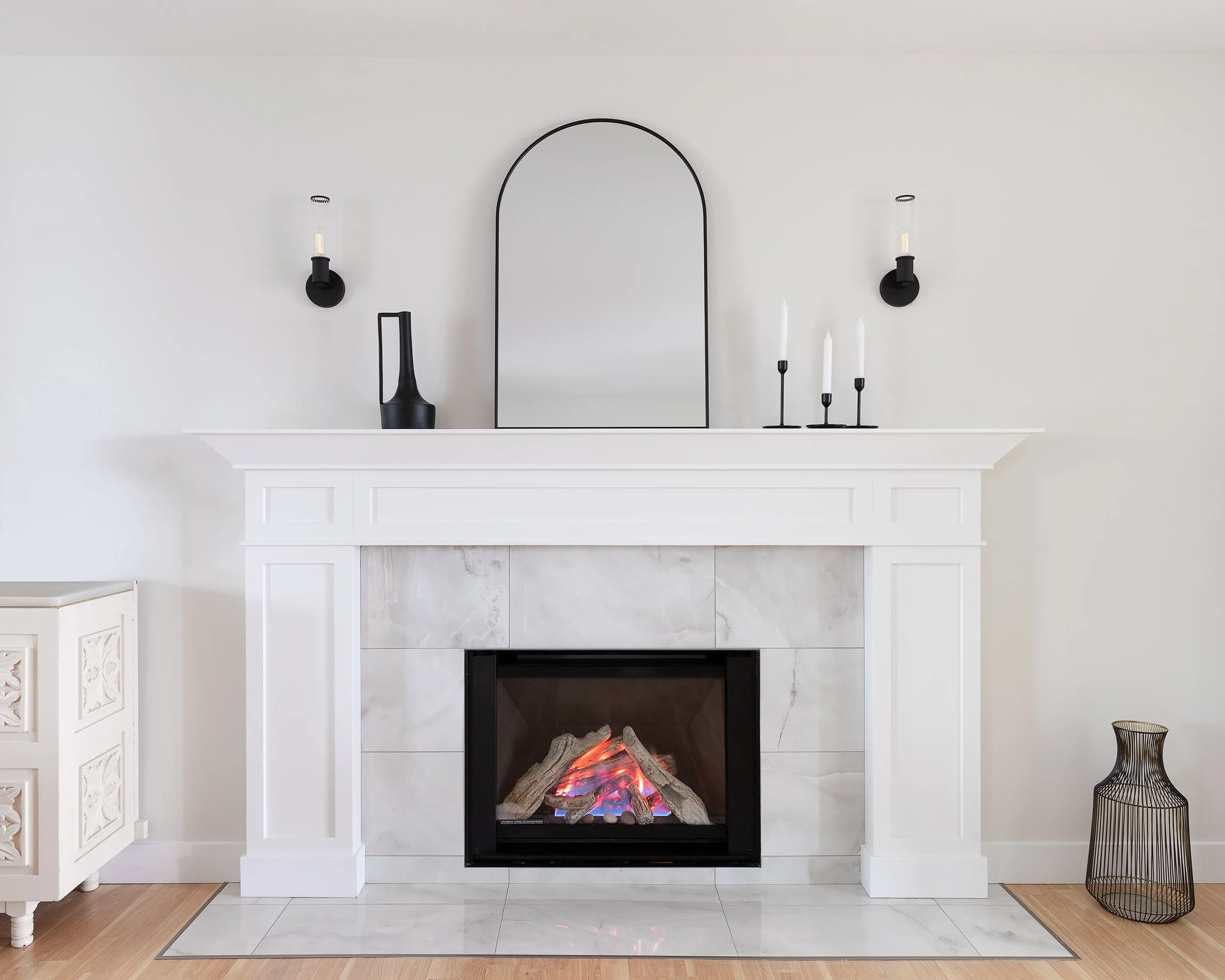 White fireplace with a black insert, a mirror and minimalist black decor on mantel, black wall sconces, and a wicker basket on wood flooring.