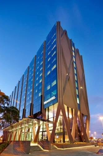 Modern high-rise hotel building with glass facade, named Hotel Grand Chancellor, with showroom or lobby visible on the ground floor, adjacent to other apartments or condos, with street and trees in front.