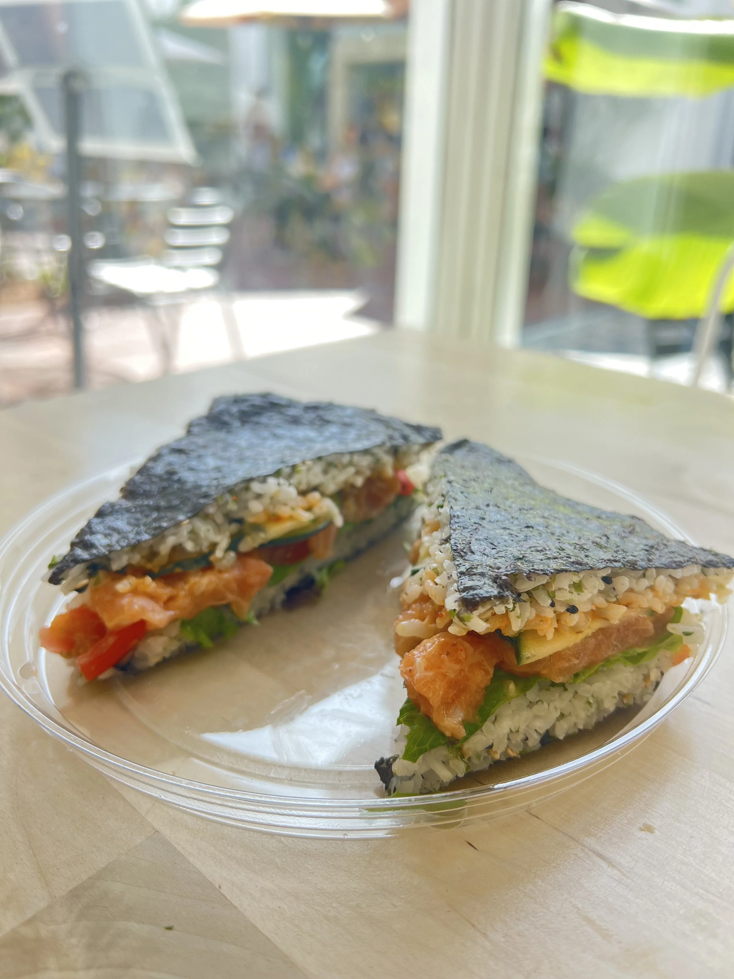 Close-up of two rice paper sushi wraps filled with shrimp, cucumber, tomato, and rice, on a clear plastic plate near a window with a blurry background.