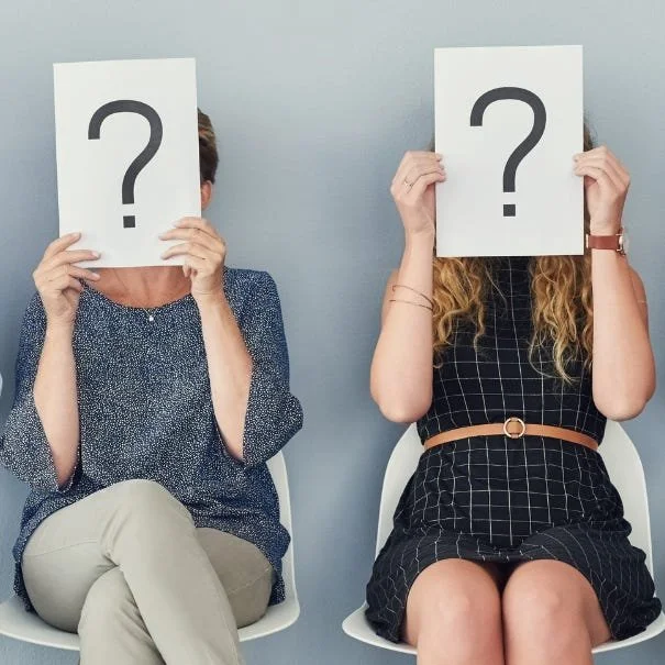 Two white women sitting in chairs. They are both holding a piece of paper with a large question mark in front of their face.