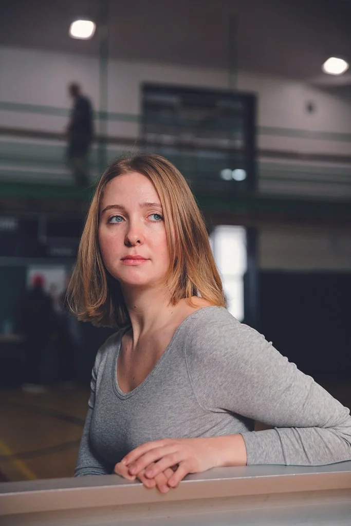 Person with shoulder-length hair wearing a gray long-sleeve shirt, standing indoors, with a blurred background of a spacious room and upper level walkway.