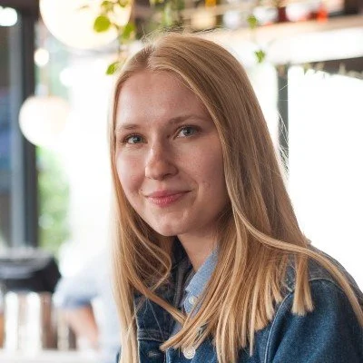Person with long blonde hair in a denim jacket smiling indoors with blurred background.