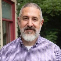 Man with gray beard, wearing a checkered shirt, standing outdoors with trees and a building in the background.