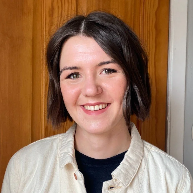Portrait of a smiling person with short brown hair, wearing a white collared shirt and black top, standing in front of a wooden background.