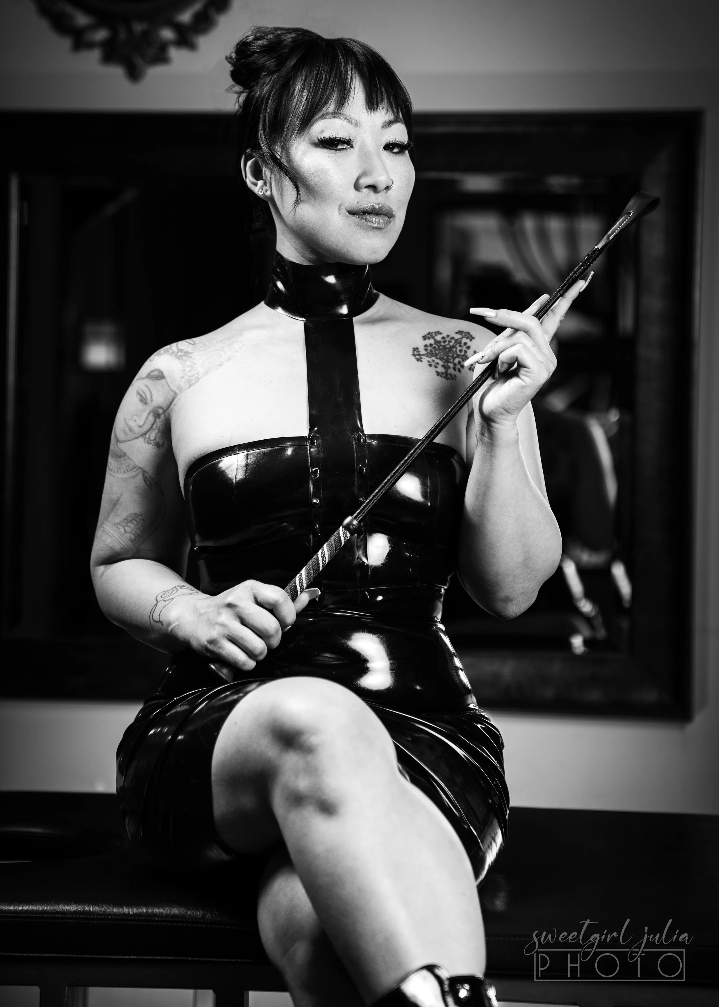 Lady Sokha, Denver dominatrix, seated in black latex corset and collar holding a riding crop
