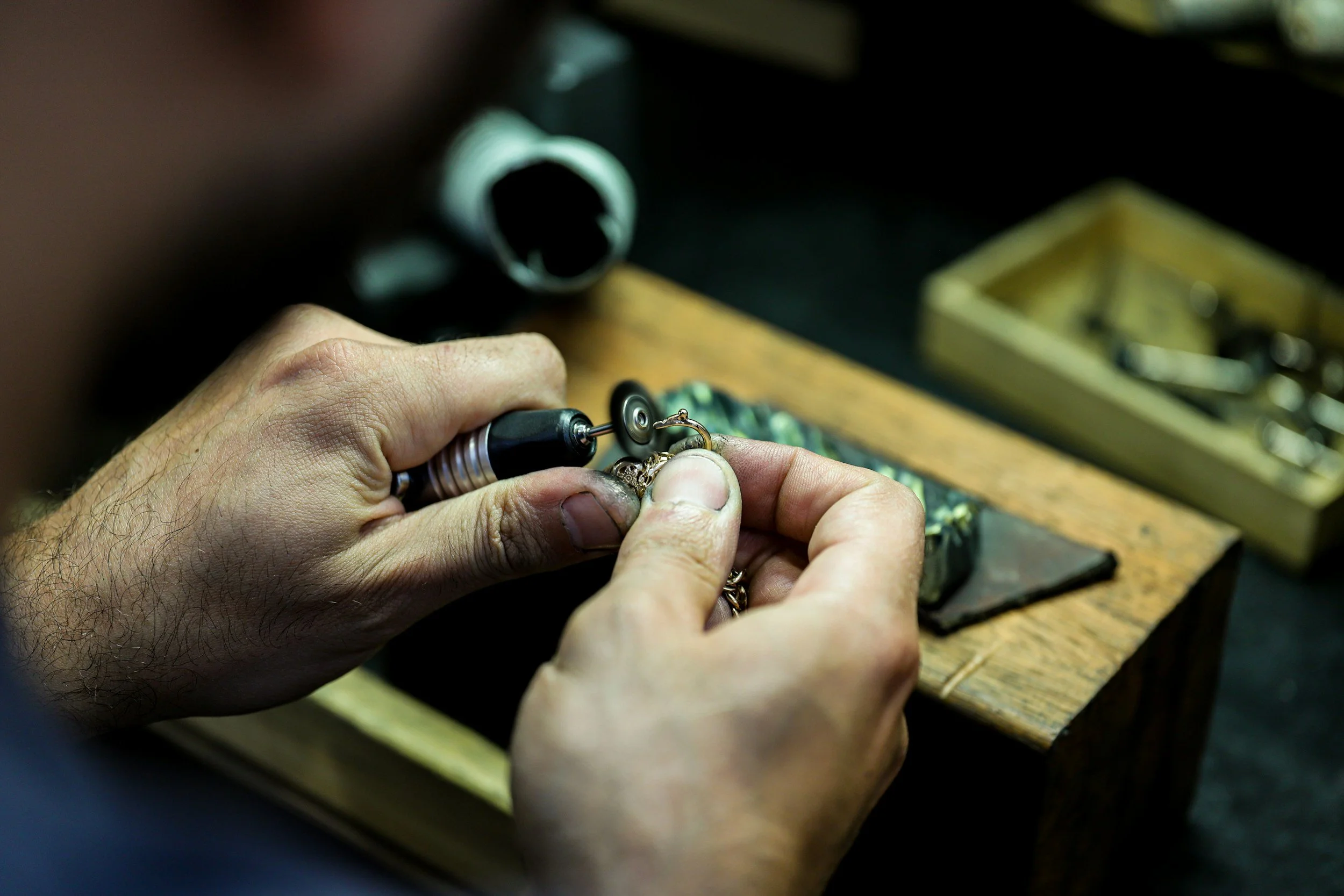 A person is working on jewelry with a rotary tool, holding a ring or piece of jewelry on a workbench in a workshop.