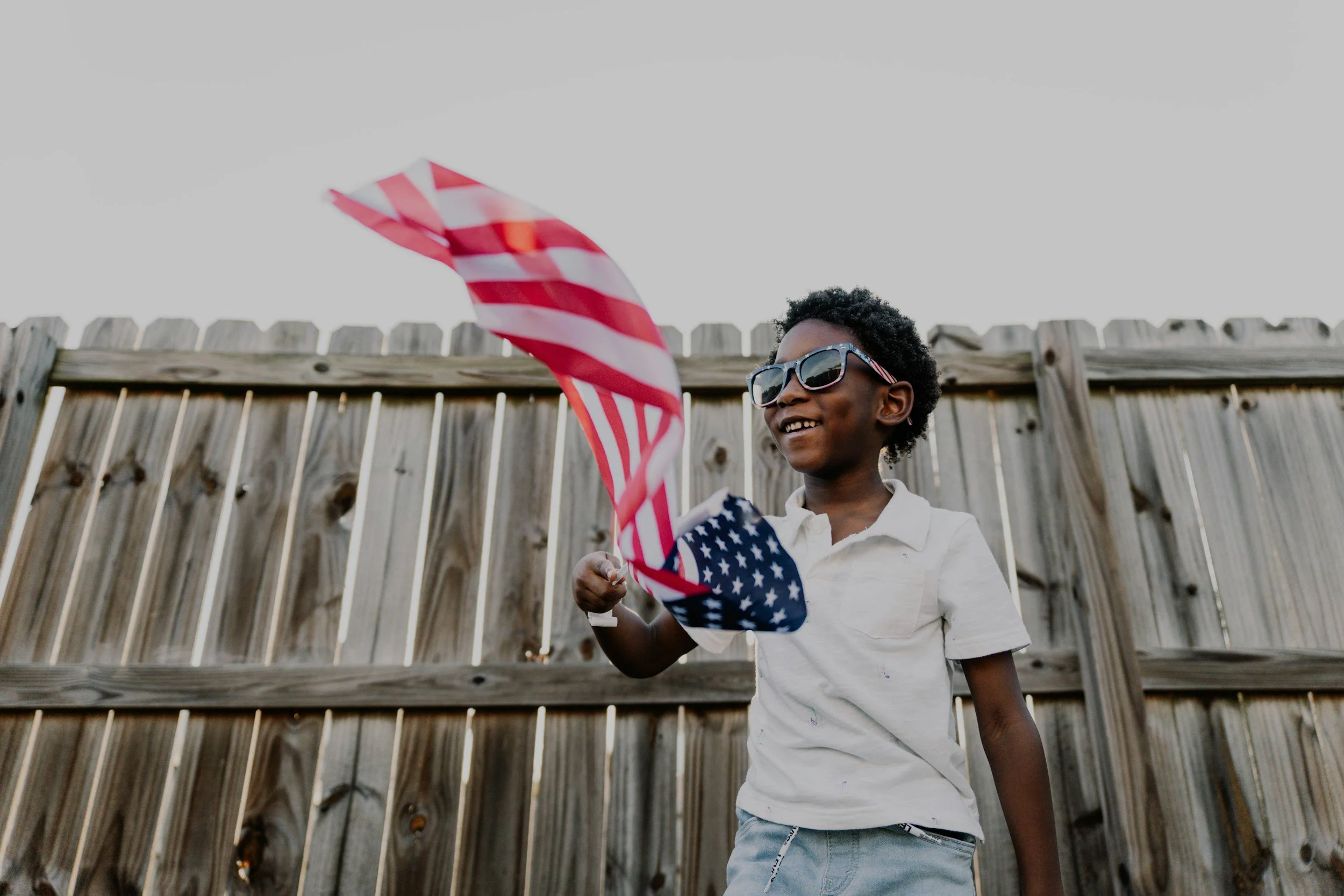A boy wearing jeans and a white polo shirt and red, white and blue sunglasses waving an American flag