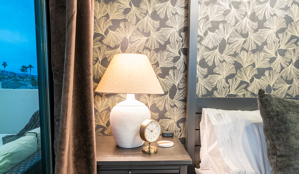 A bedside table at Vista Lago with a beautiful lamp and clock and stunning wallpaper as a backdrop