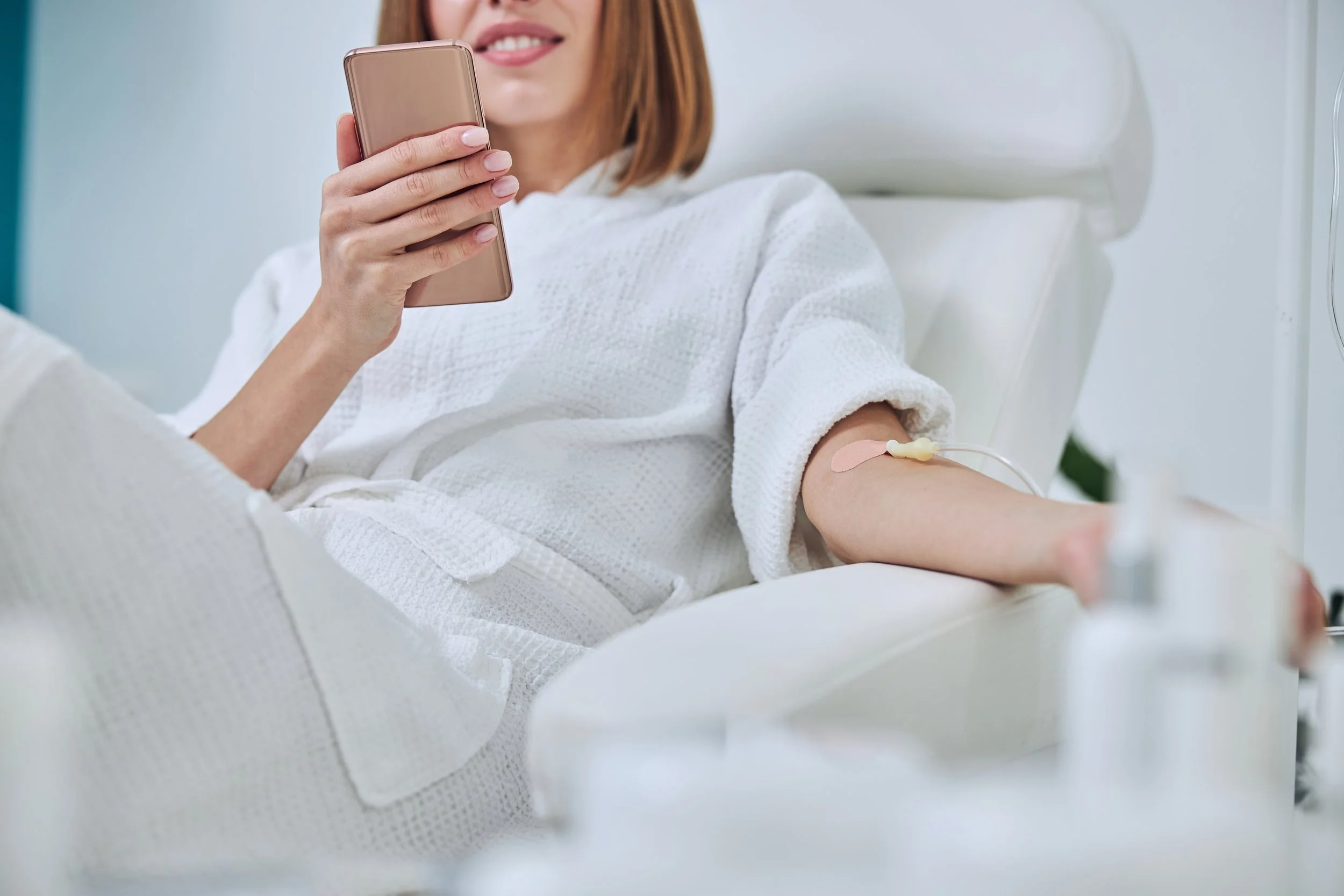 Woman in a white robe having an IV therapy while holding a smartphone.
