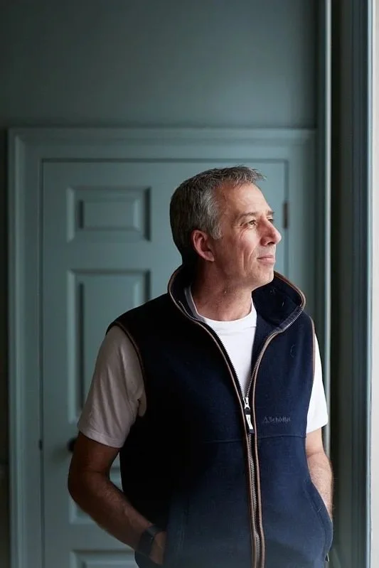 An older man with gray hair stands by a window, gazing outside with a thoughtful expression, wearing a white T-shirt and a navy blue vest.