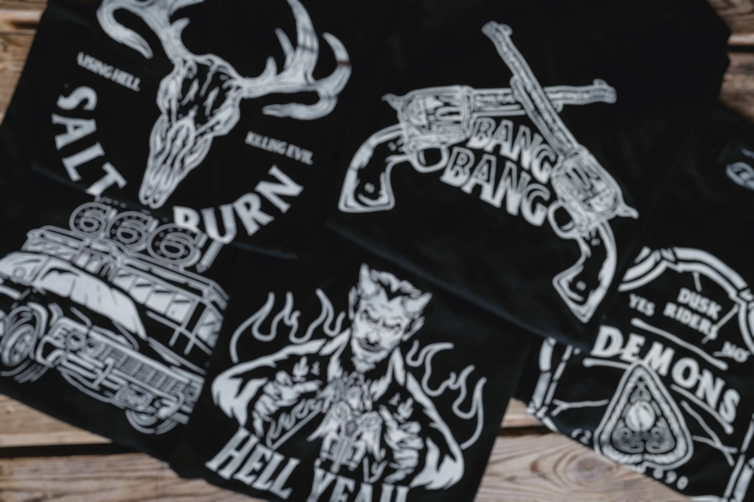 Black cloth with white graphic designs of a galloping horse, a revolver with 'BANG BANG' written, a devil with flames and 'HELL YES' text, a vintage truck, and phrases like 'DURAN', 'SALTY', 'DEMOMS', and 'DUSK RIDEY' printed in bold letters across the cloth.