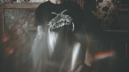 A person in a black t-shirt with a graphic design, seated indoors with animal print and floral patterned walls in the background, featuring denim shorts.