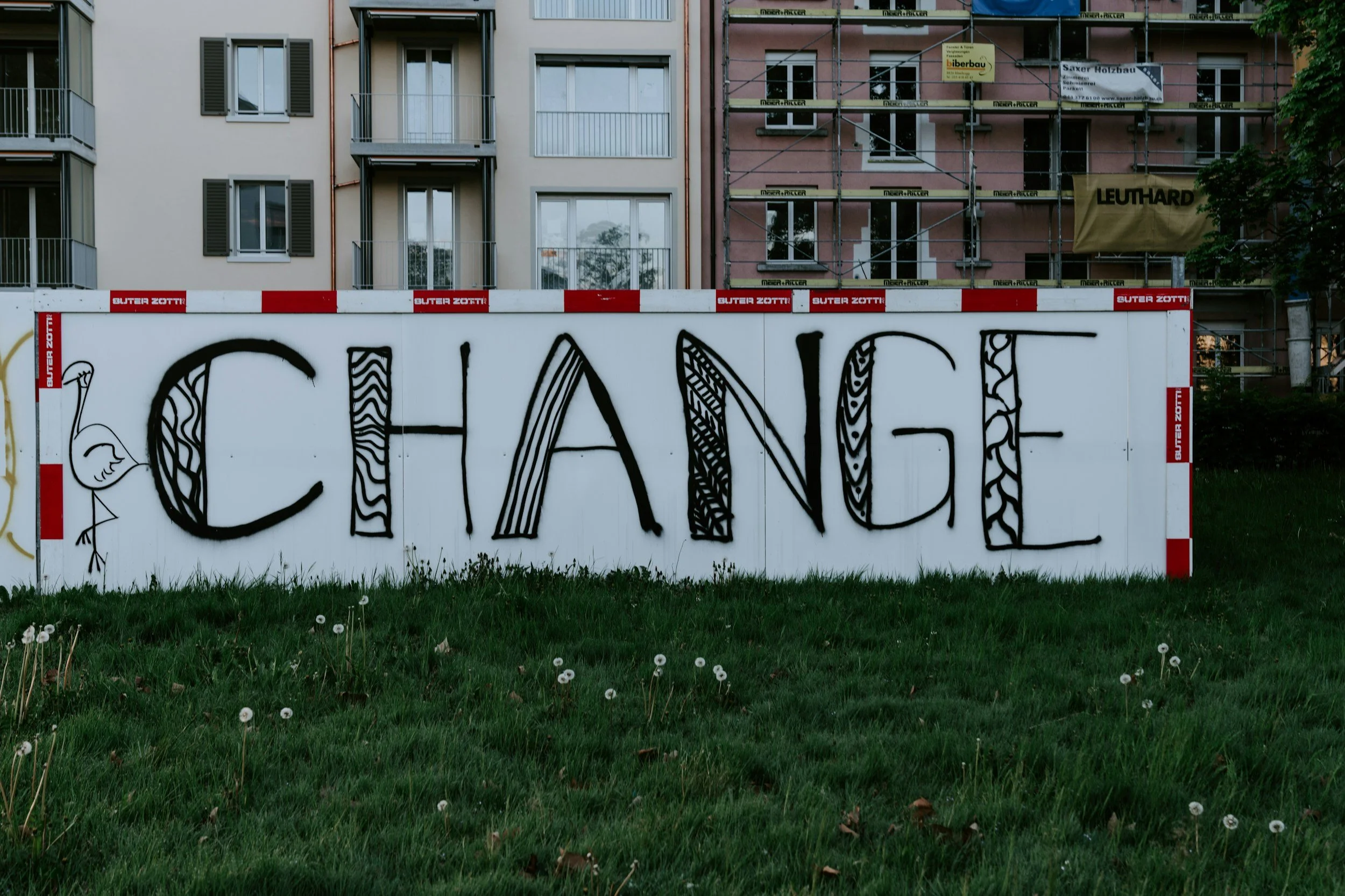 A large white sign with the word 'CHANGE' spray-painted in black, decorated with various patterns within each letter, is positioned in front of a grassy area with dandelions. In the background, there are residential buildings, some under construction with scaffolding.