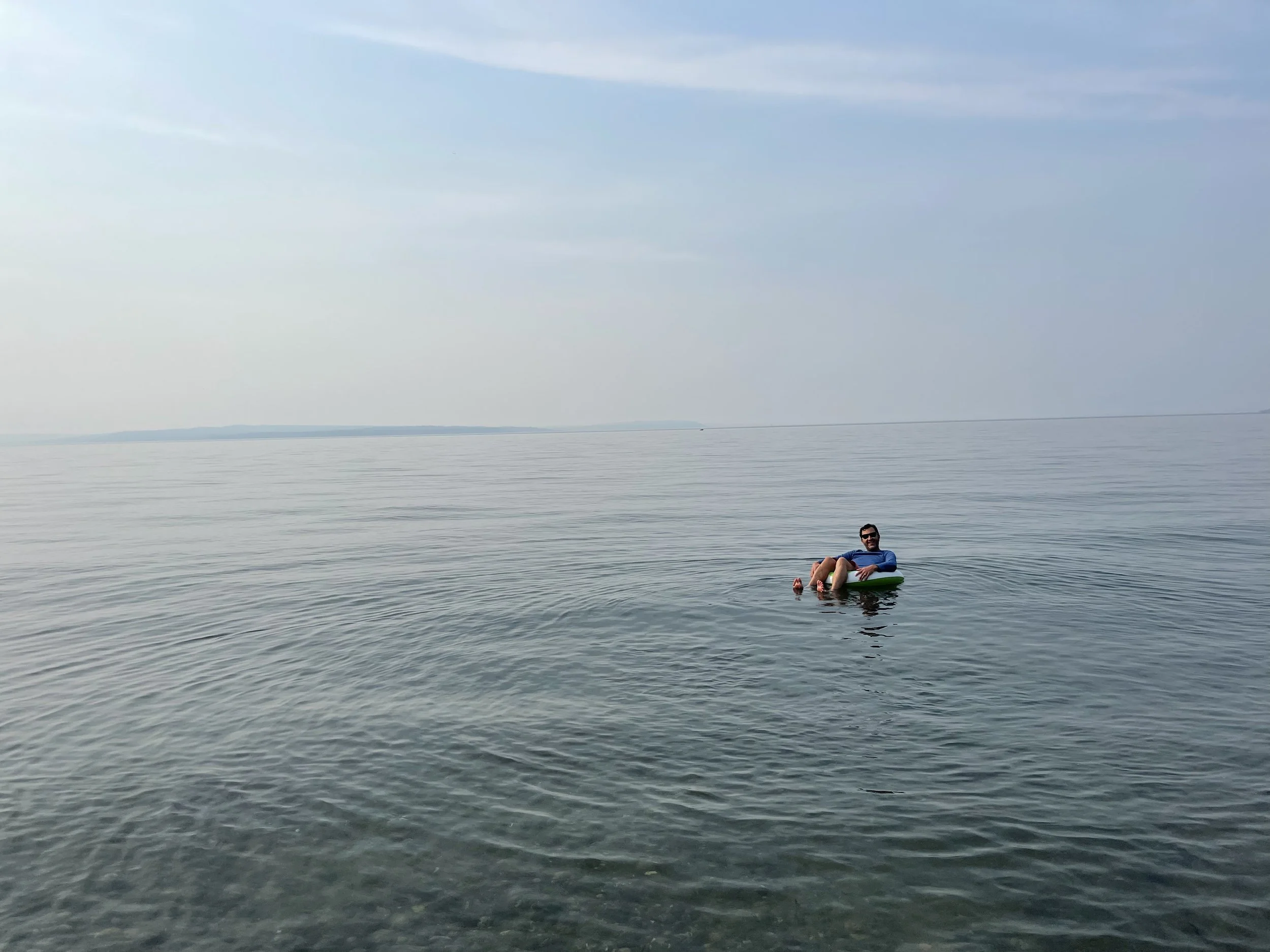 my husband Jesse, floating on a floaty in Puget Sound off north beach in summer.  The camera is zoomed out and it's a hazy day, so most of the image is water and sky, and it looks like he's drifting away.  He has a silly grin on his face.