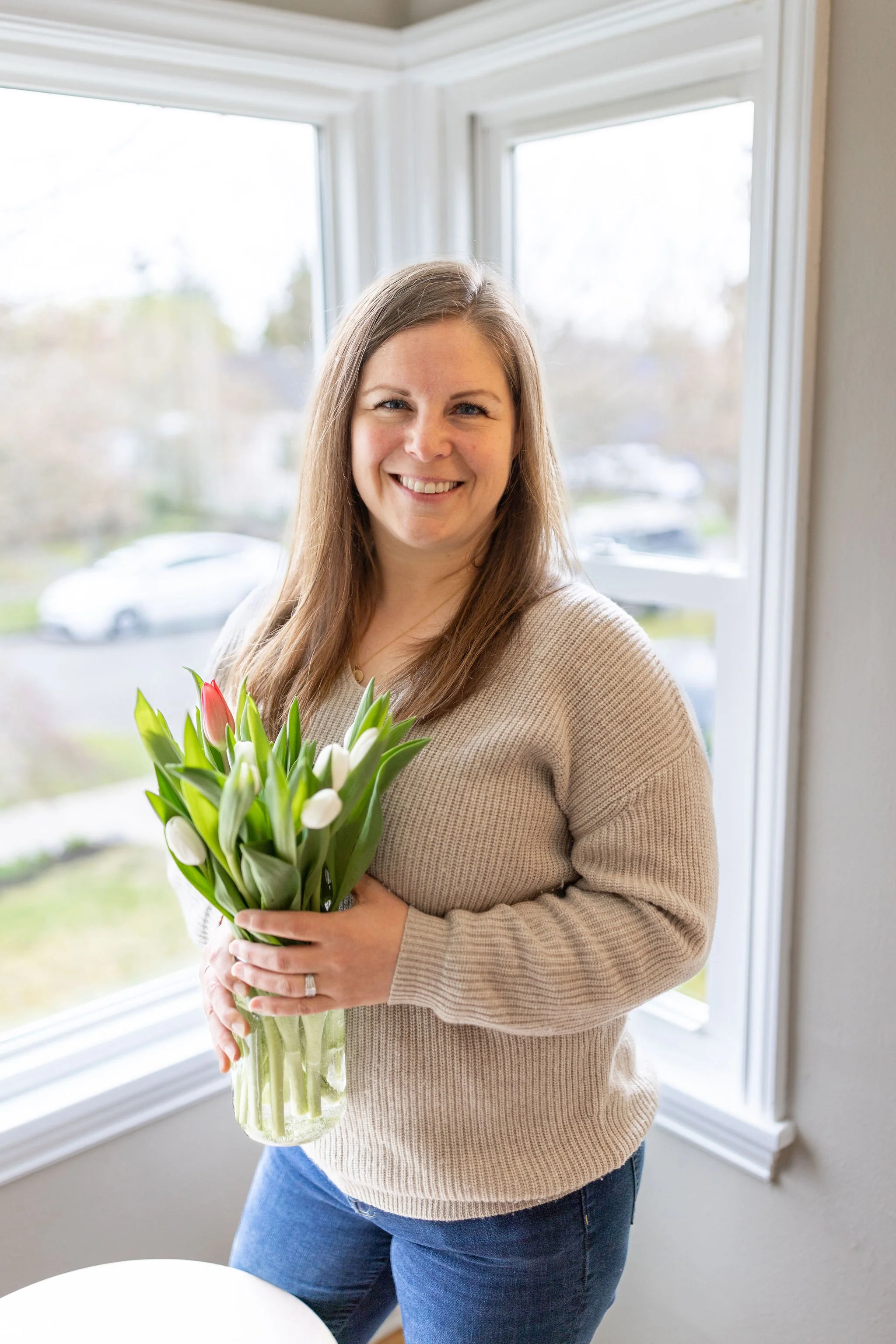 Peri standing in front of a window in one of her listings, smiling, and holding a vase of tulips that will be part of her staging.