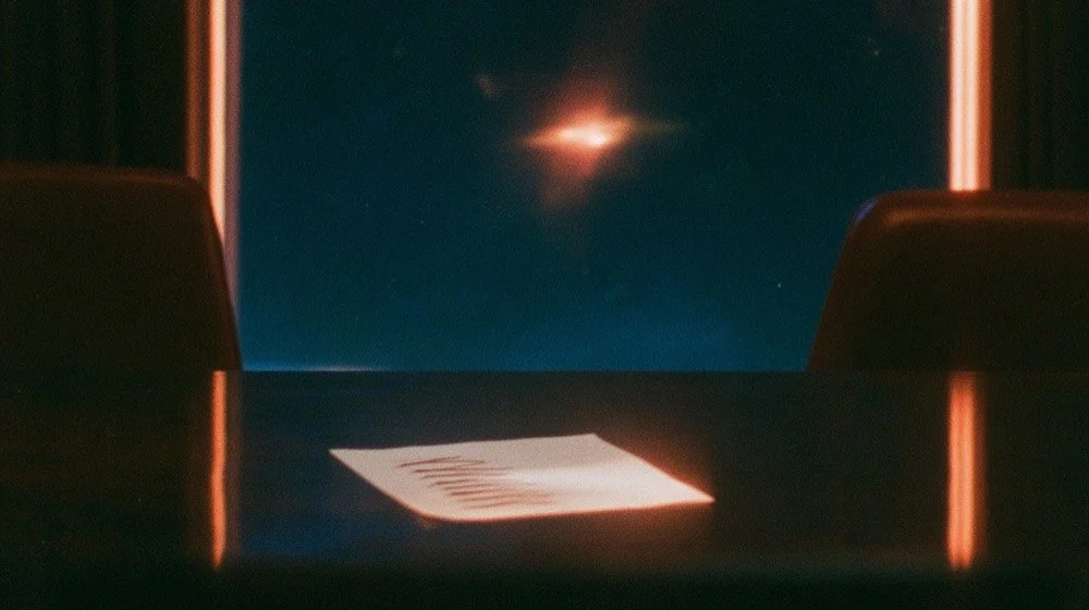 A single handwritten sheet of paper on a dark conference table between two chairs, with a bright star of light flaring in the window behind.