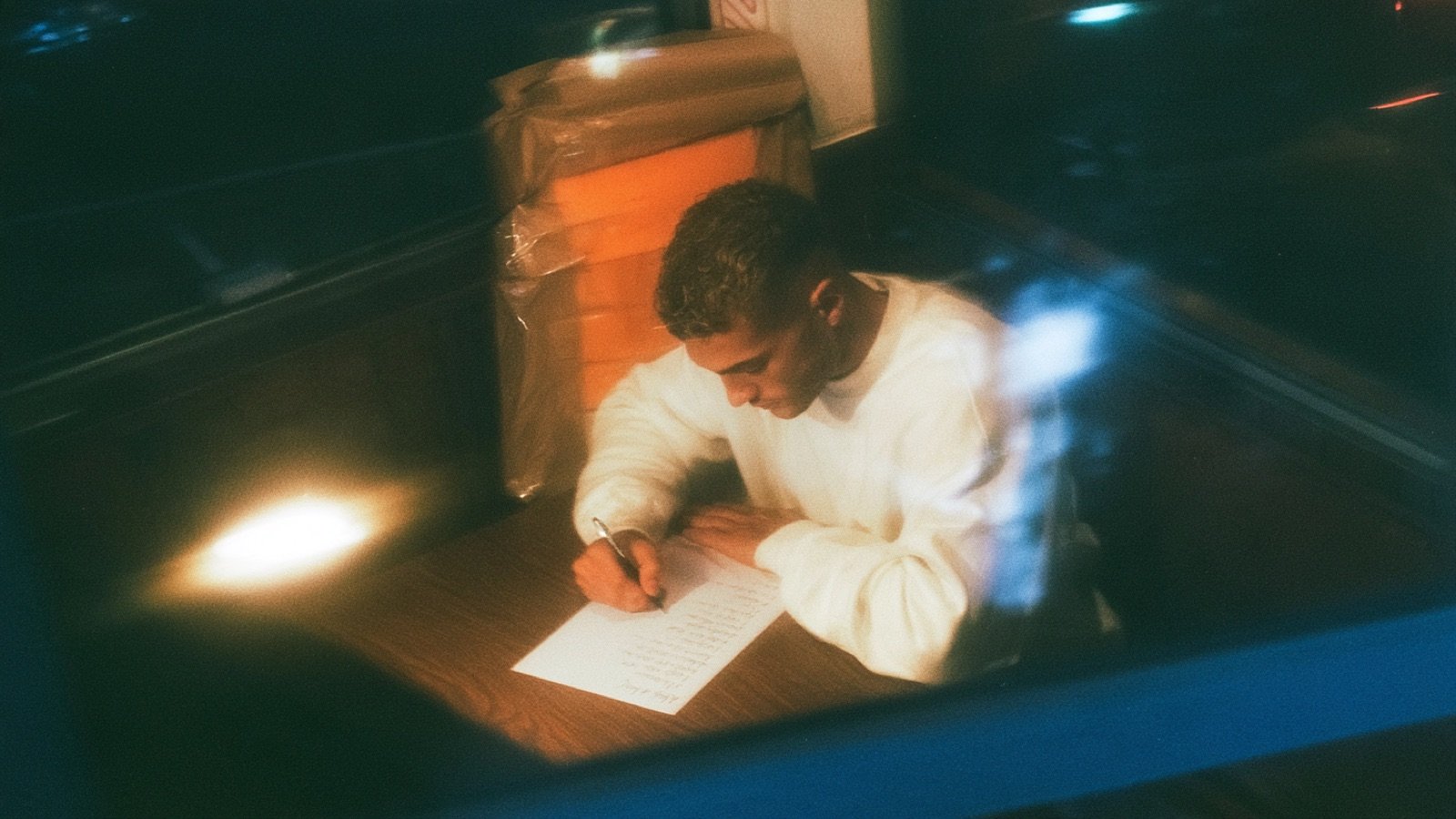 A young man in a white sweater writes a list by hand on a single sheet of paper at a wooden table, seen at night through a window.